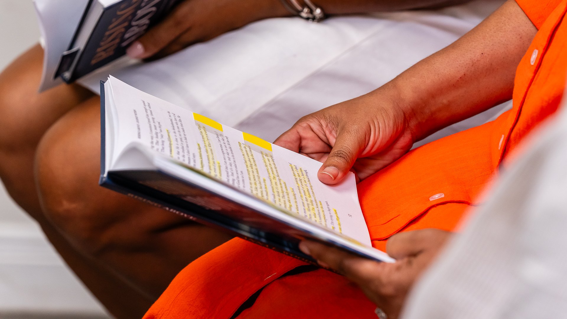 Close-up of a person reading a magazine with highlighted text, sitting with their legs crossed and wearing bright orange shorts.