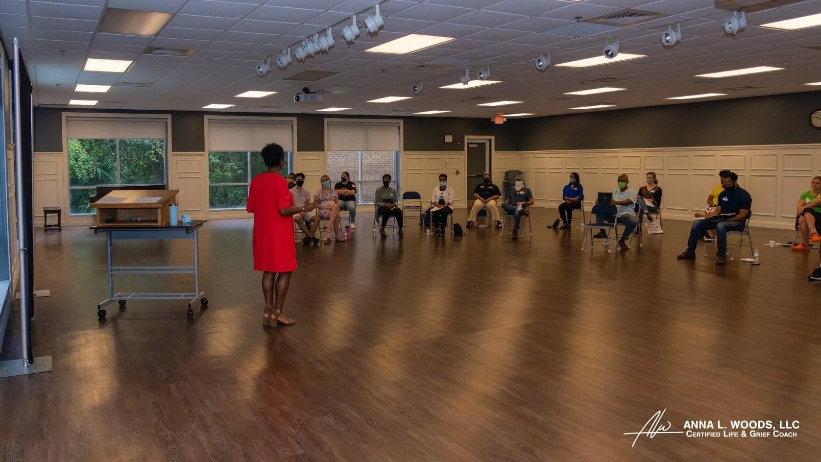 A woman in a red dress standing and speaking to a seated audience in a large, well-lit room with wood flooring and windows. The audience members are wearing masks and sitting apart.