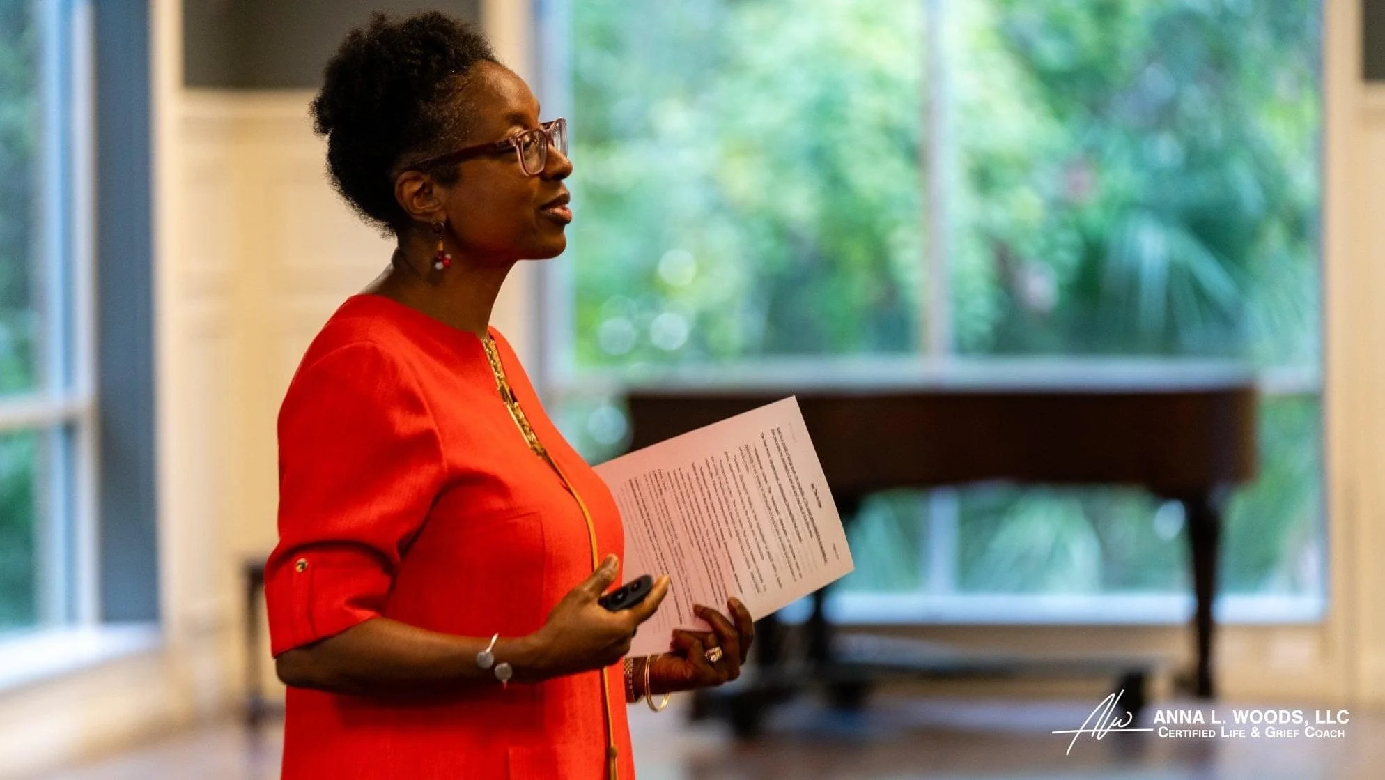 A woman with short curly hair, wearing glasses and a red dress, standing indoors near a large window, holding a paper and a remote control. A grand piano is visible in the background.