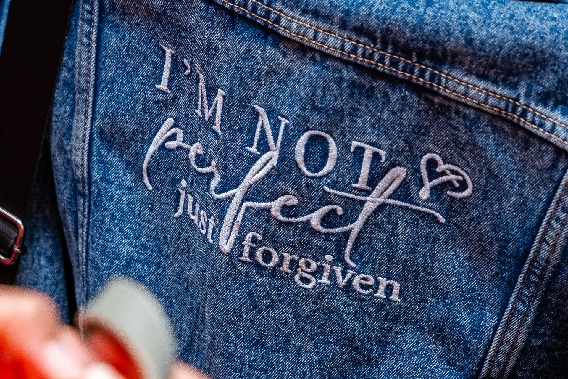 Close-up of a person's denim jacket with embroidered text reading 'I'M NOT perfect but I am love.' in white thread.
