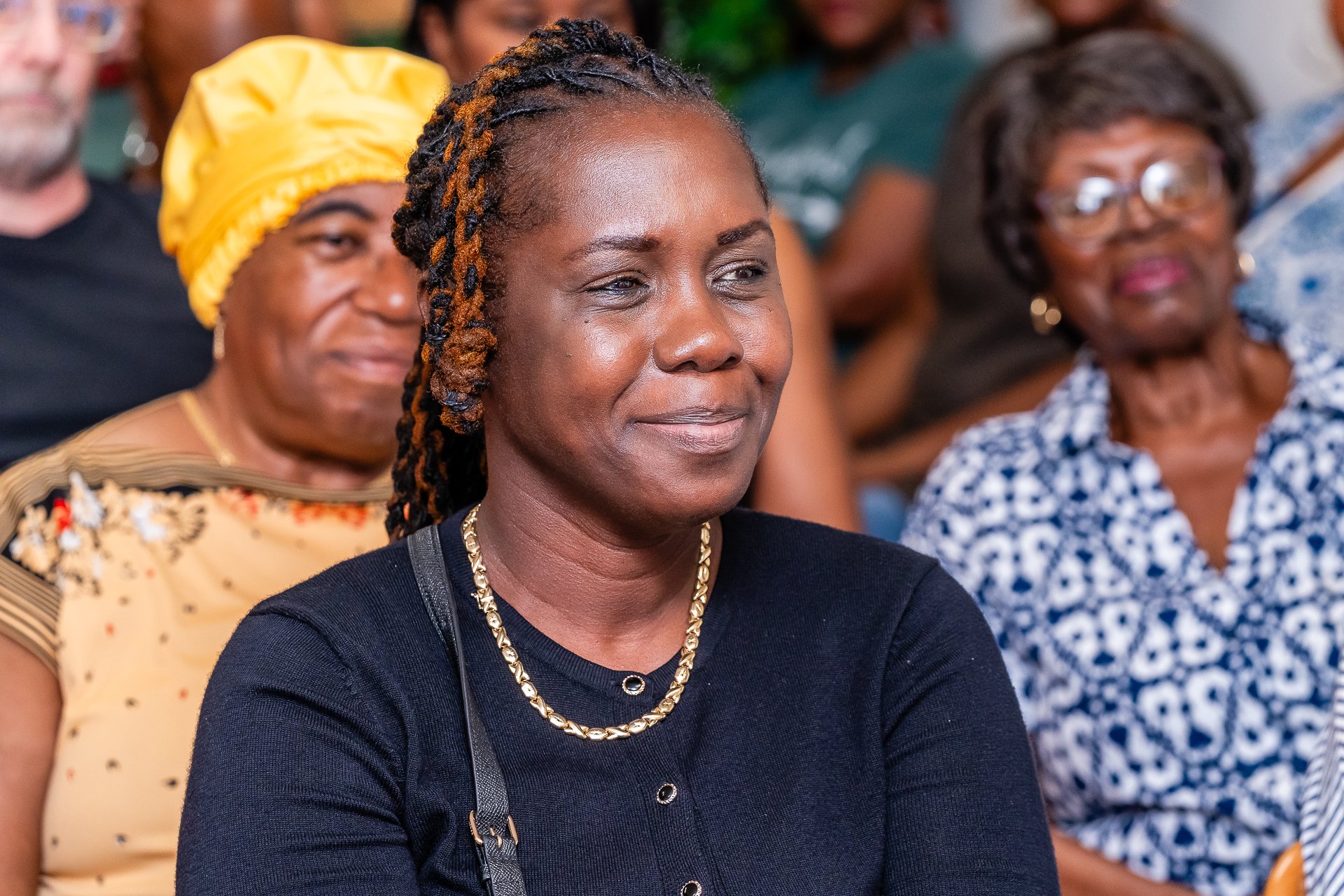 A woman with braids and a gold chain necklace is attending an event, sitting among diverse women in the background.