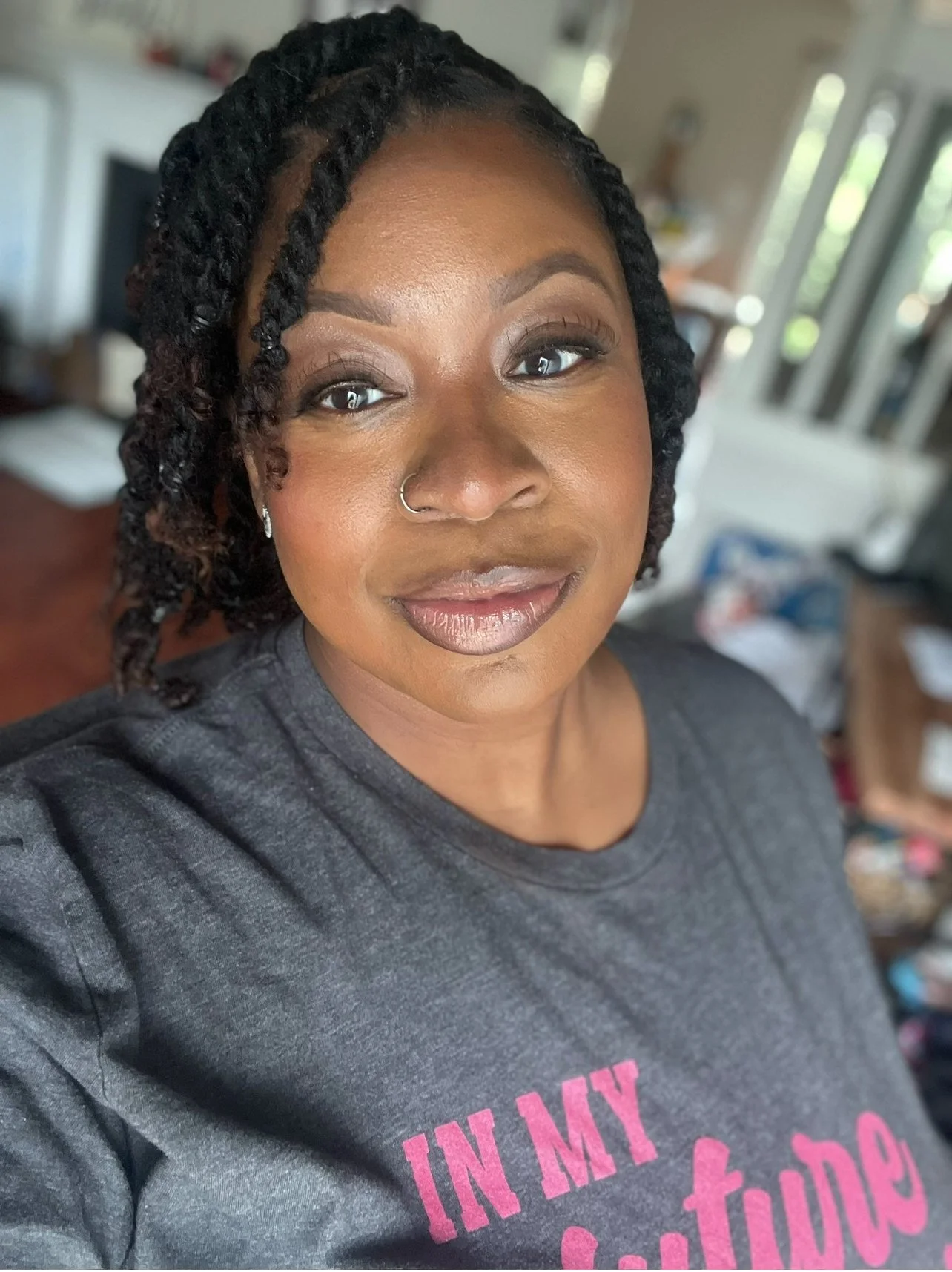 A woman with braided hair, wearing makeup, a nose ring, earrings, and a gray t-shirt that says "IN MY" in pink letters. She is smiling slightly and standing in a room with natural light.