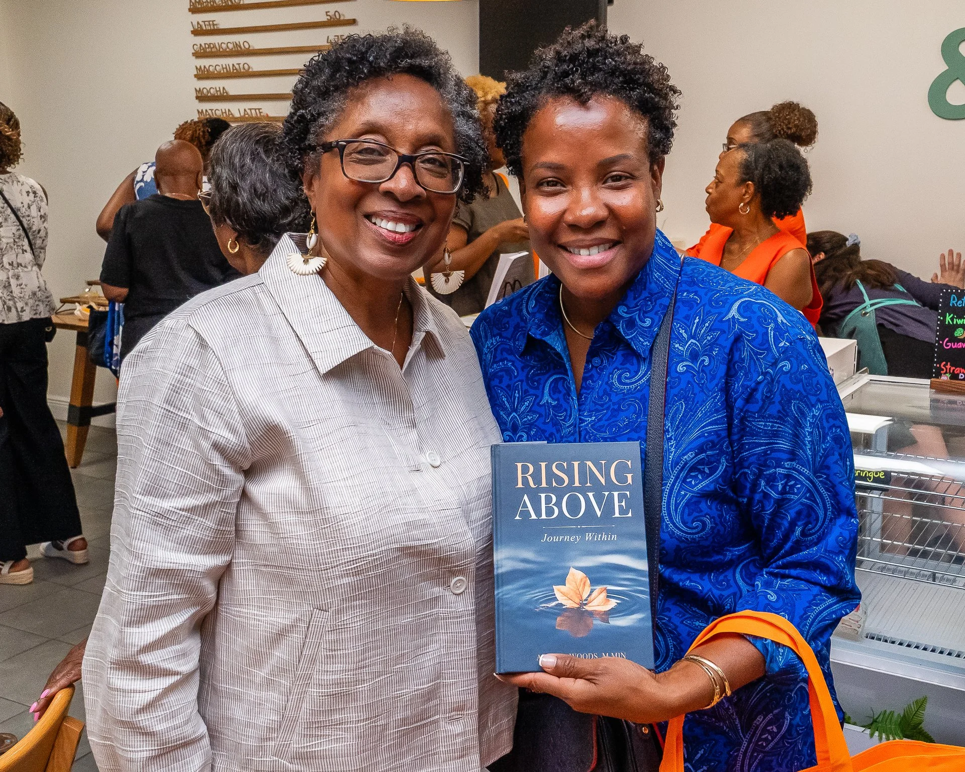Two women smiling at the camera, one wearing glasses and earrings, the other holding a book titled "Rising Above" in a bookstore or library, with other people and bookshelves in the background.