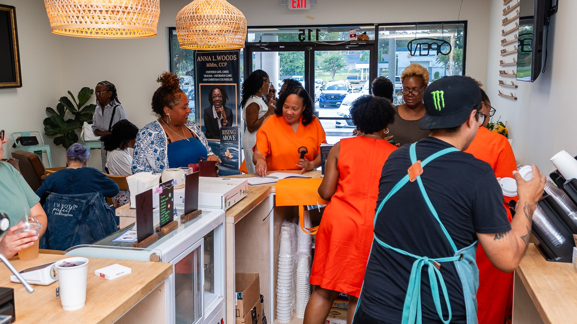 People gathered inside a store, some wearing orange aprons, engaging in conversations, with a woman at the counter reading a book, and various products displayed on the counter.