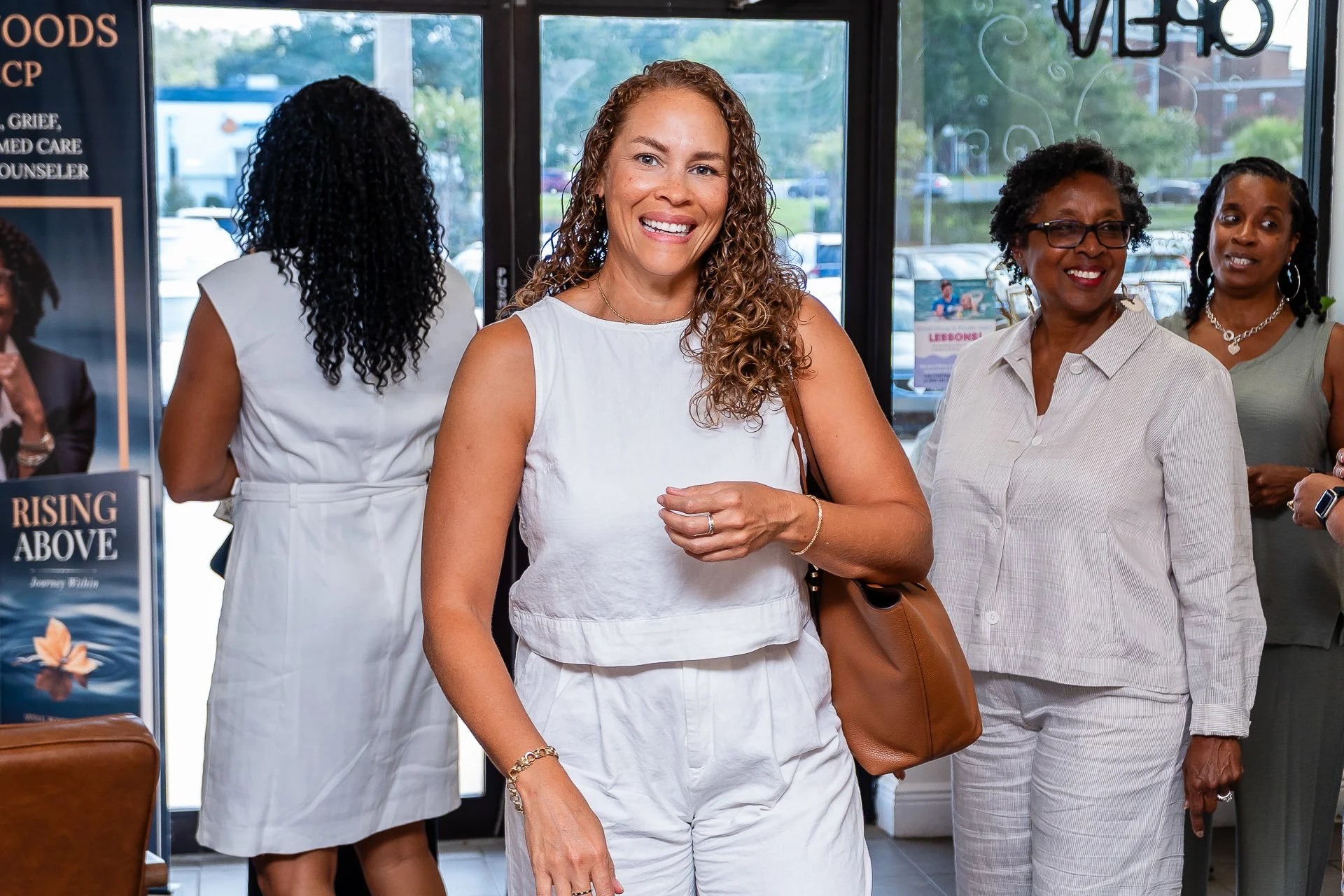 Group of women standing inside a building near large glass doors, smiling, with books and posters visible in the background.