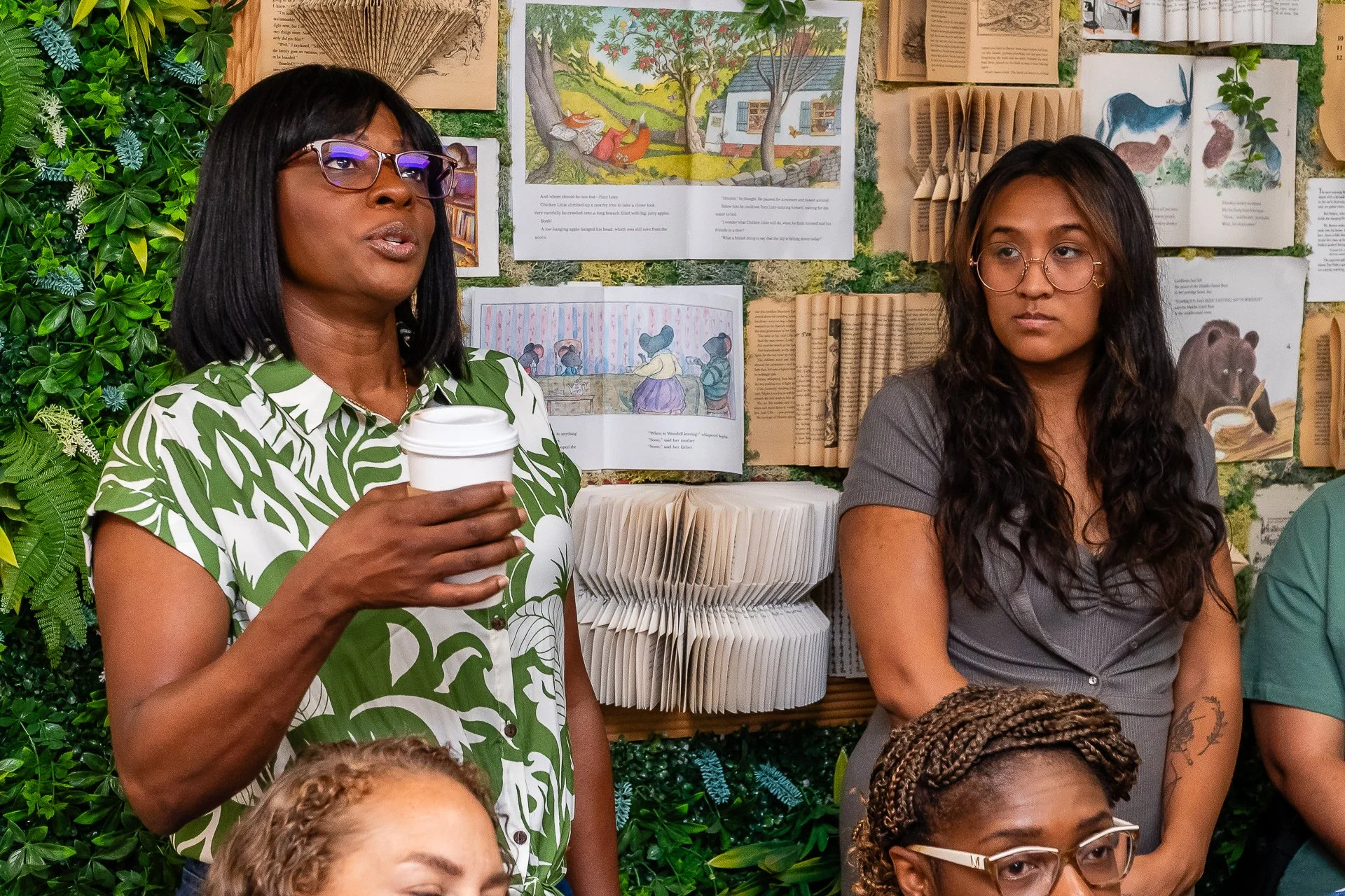 Two women are sitting in front of a display of artwork and open books. The woman on the left, wearing glasses and a green patterned shirt, is holding a disposable cup. The woman on the right, also wearing glasses, has long, dark hair and is wearing a