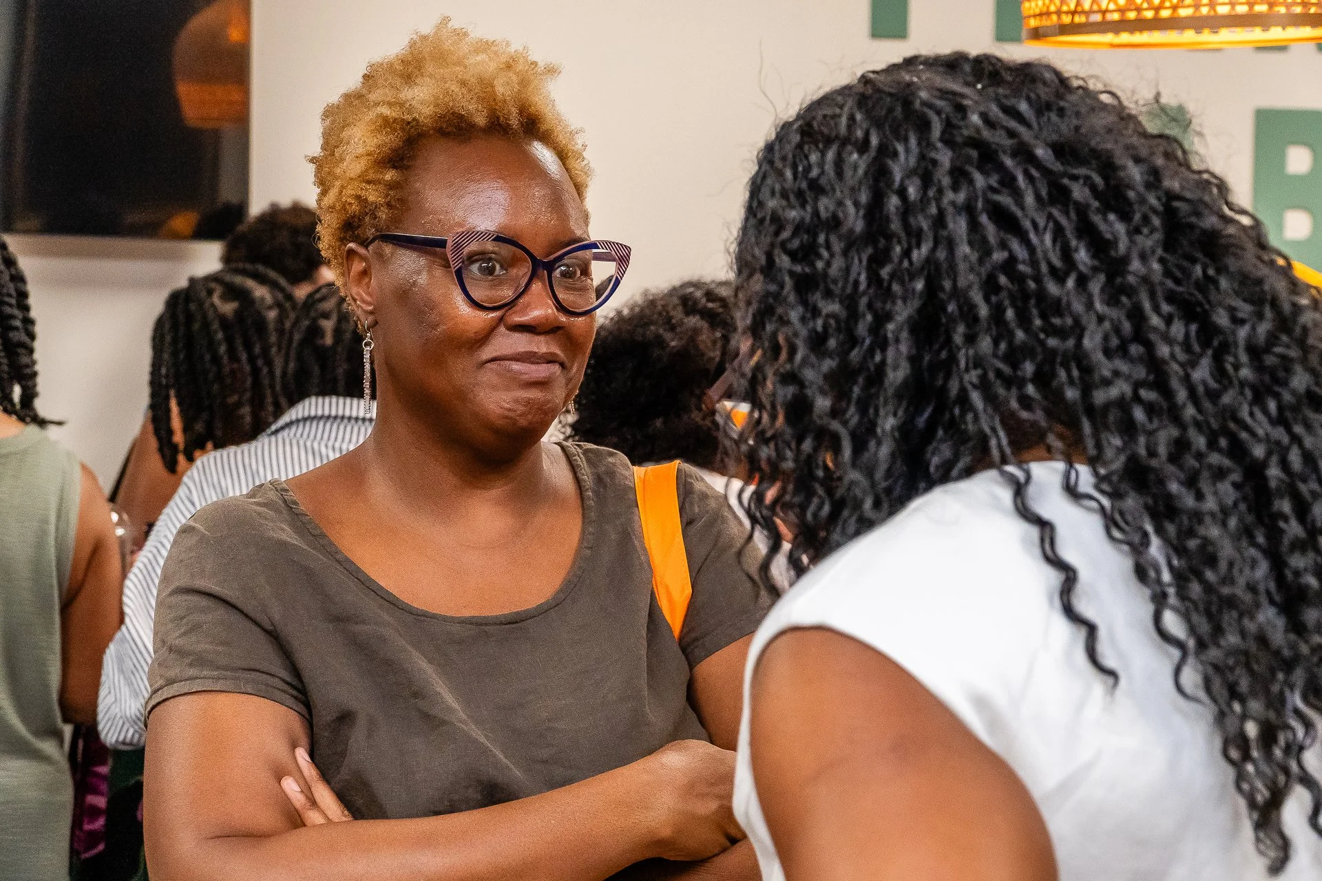 Two women are engaged in a conversation indoors, one woman with short blonde hair, glasses, and a gray shirt, facing the other woman with long curly black hair and a white shirt. Several other people are blurred in the background.