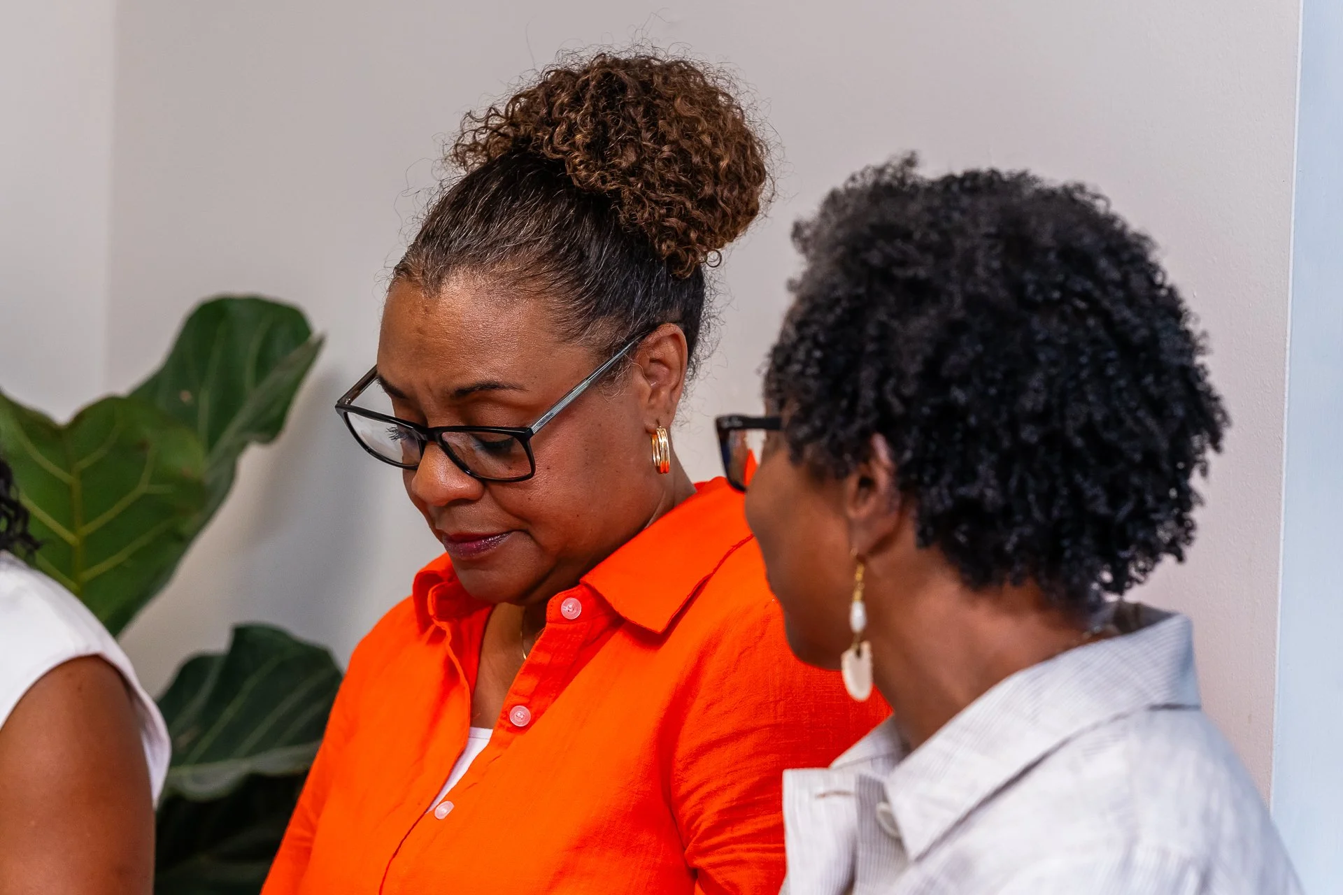 Two women in conversation, one wearing glasses and an orange blouse, the other with curly hair and earrings, indoors with a plant in the background.