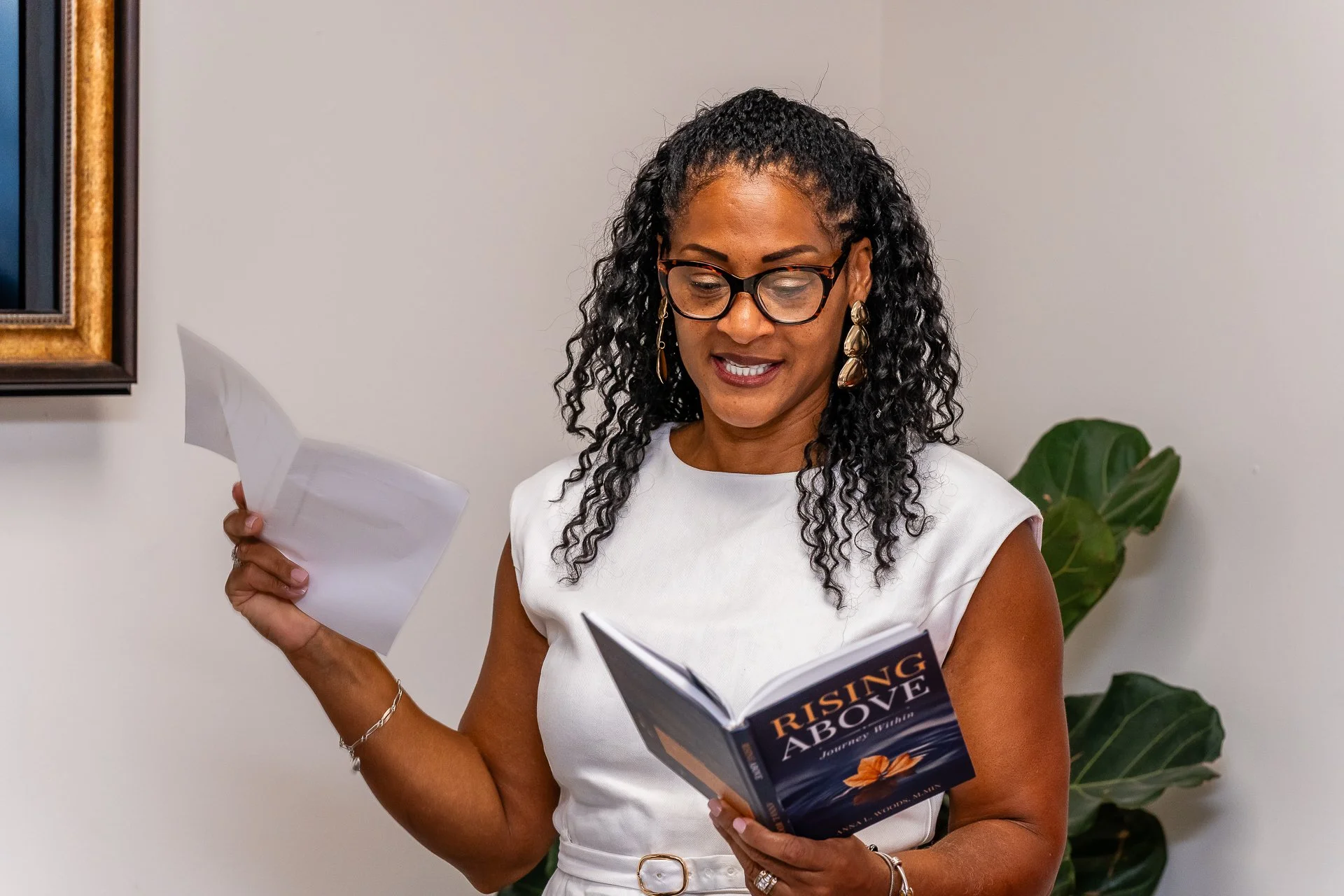 Woman with curly black hair and glasses reading a book titled 'Rising Above' while holding papers in a room with a plant and artwork on the wall.