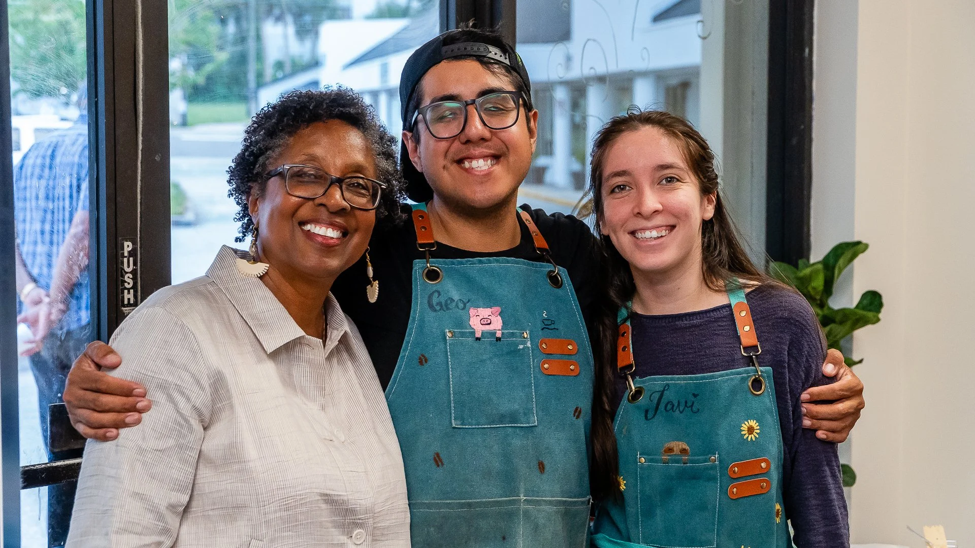 Three smiling people with aprons, hugging inside a store or café, near a door with a "Push" sign, with a woman on the left, a man in the middle, and a woman with a name tag that says 'Javi' on the right.
