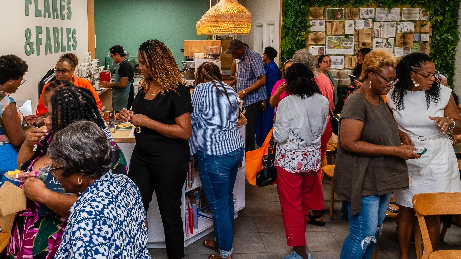 People standing in line at a coffee shop or cafe, some holding phones or eating, with a green wall and a sign that reads 'FLAKES & FABLES' in the background.