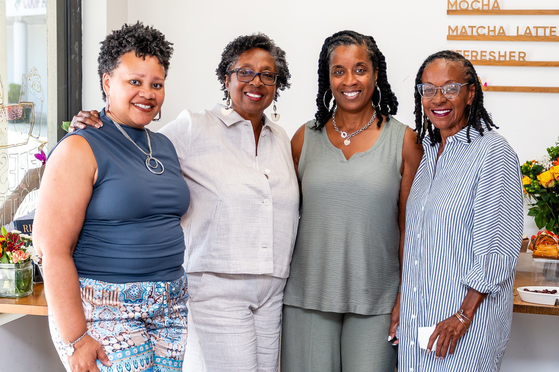 Four women of African descent standing together indoors, smiling at the camera. Behind them is a menu board with drinks such as mocha, matcha latte, and refresher, and a table with flowers and baked goods.