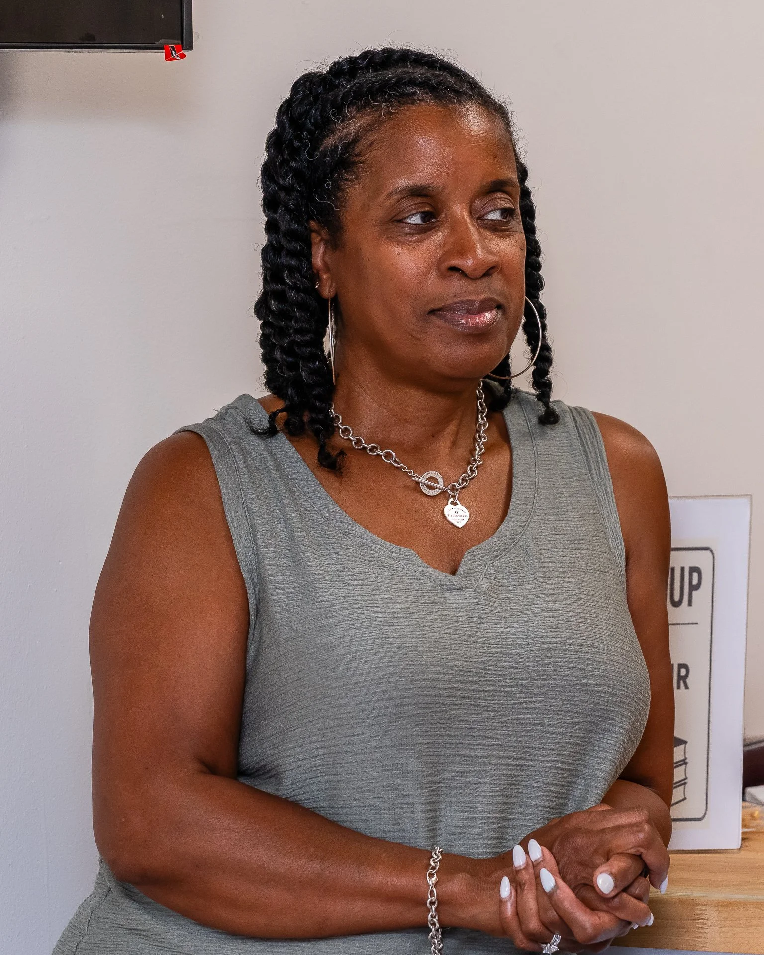 A woman with dark, curly hair styled in twists, wearing a sleeveless gray top, silver jewelry including hoop earrings, a necklace, and a bracelet, standing indoors against a plain white wall with a partially visible sign and a panel on the wall.