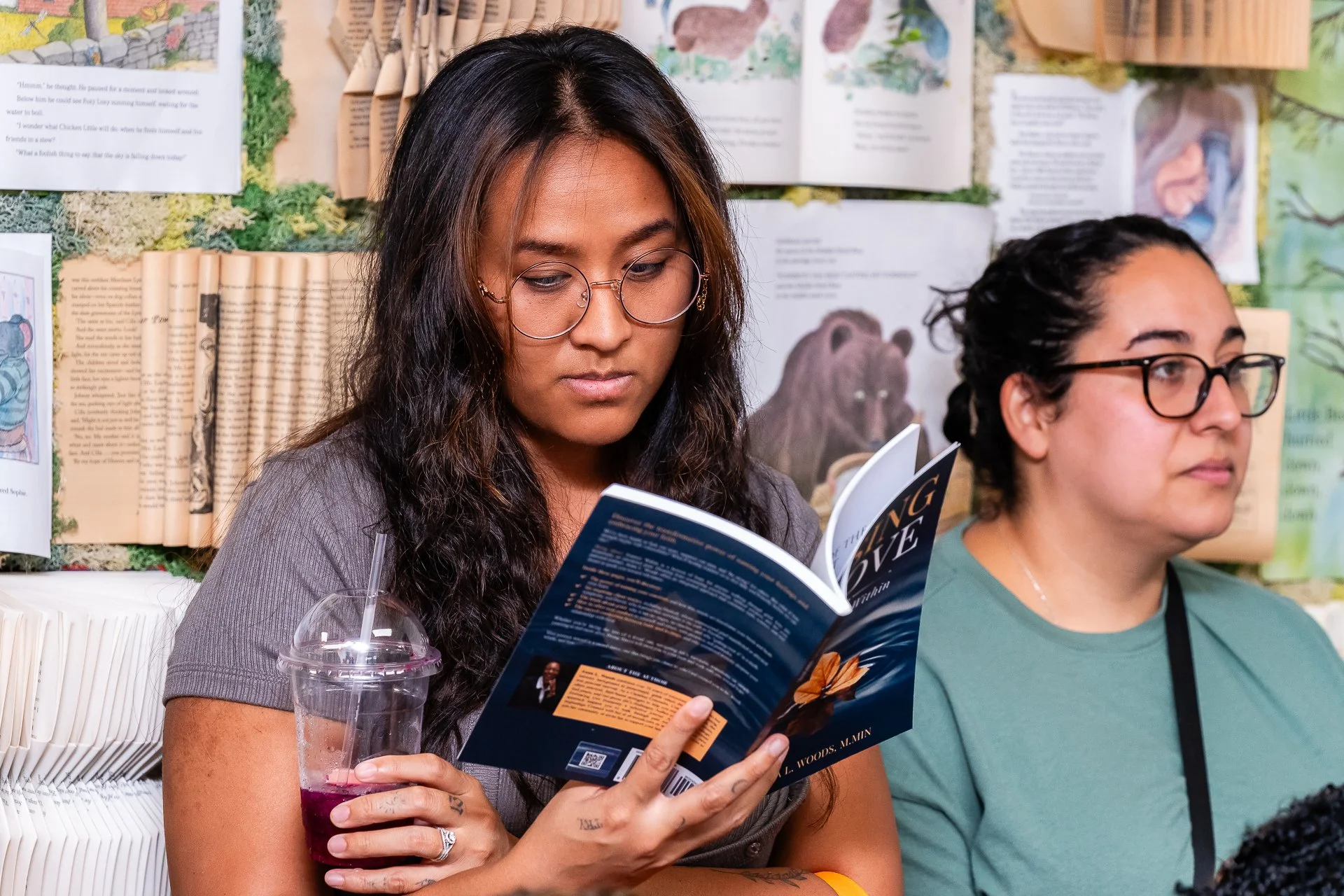 Two women sitting indoors, one reading a book titled 'The Long Goodbye' and holding a drink with a straw, the other sitting beside her. Background includes bookshelves and informational posters.