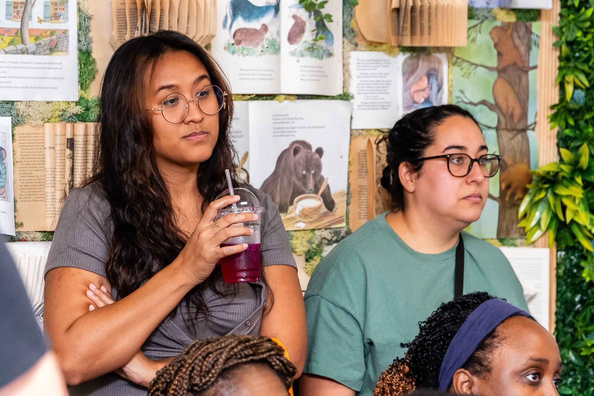 A group of diverse people attending an event, with a woman in glasses holding a purple drink and a man in glasses standing next to her. The background features educational or informational posters and a decorative plant.