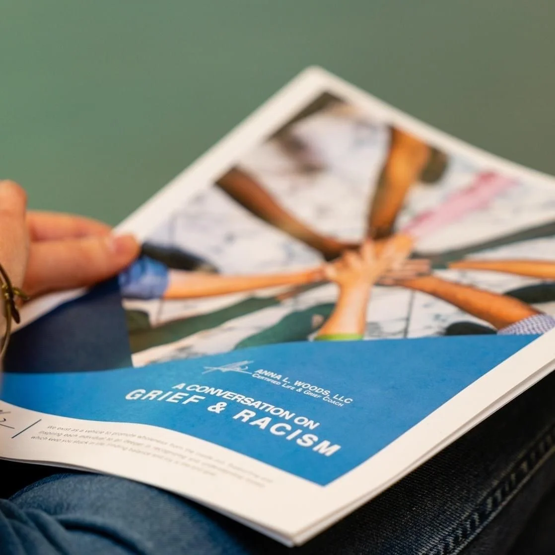 Close-up of a person holding a pamphlet about grief and racism, with a blue section and text that reads "A Conversation on Grief & Racism."