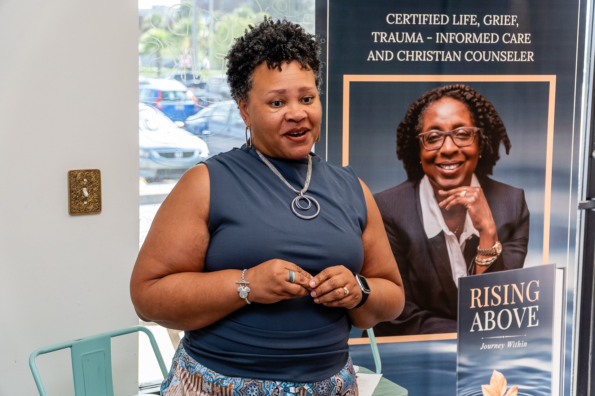 A woman with short, curly black hair wearing a sleeveless blue top, silver jewelry, and a smartwatch, standing in front of a poster about grief and trauma recovery.