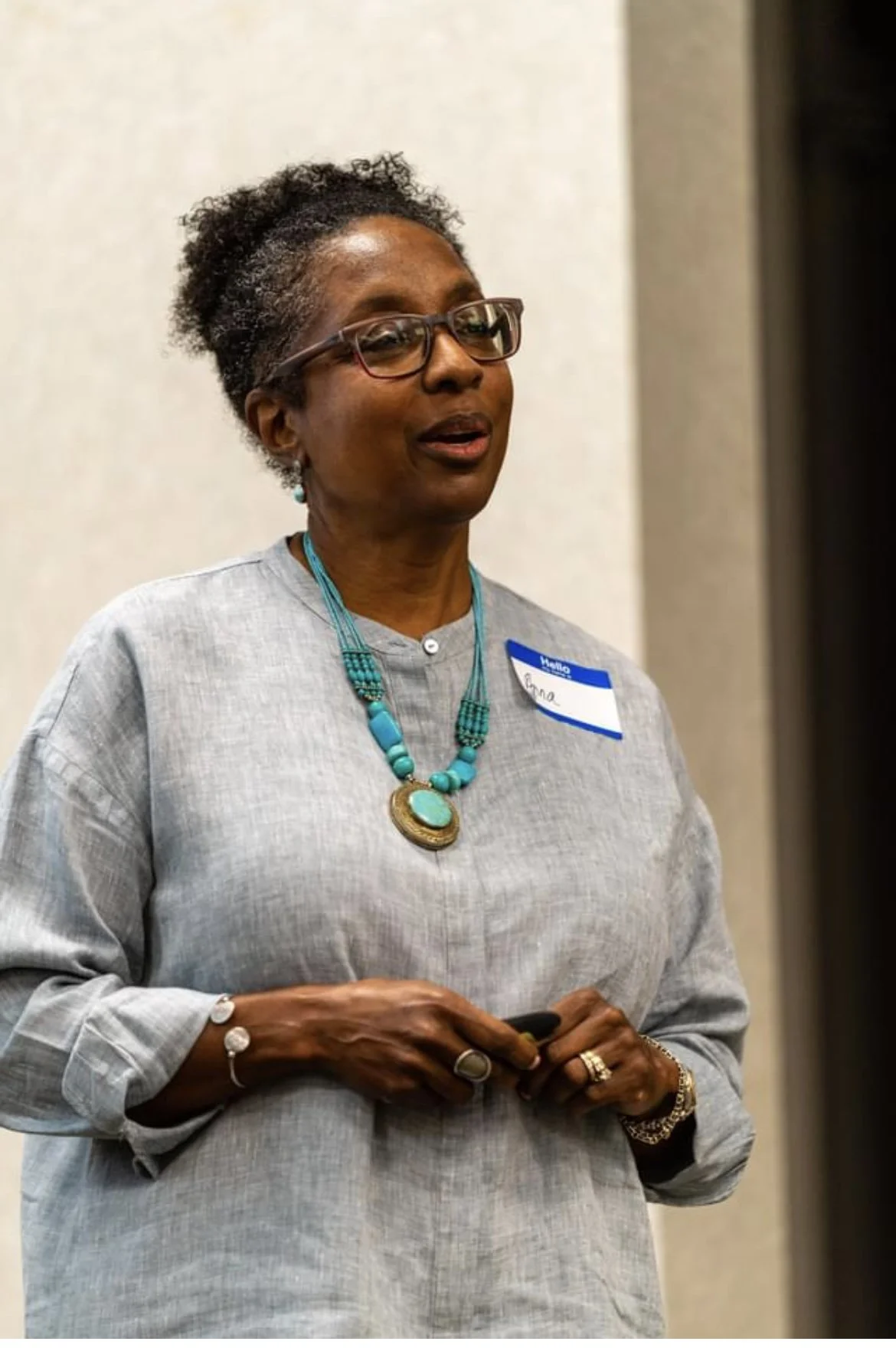 A woman with glasses and jewelry speaking at an event, wearing a gray top and a name tag.