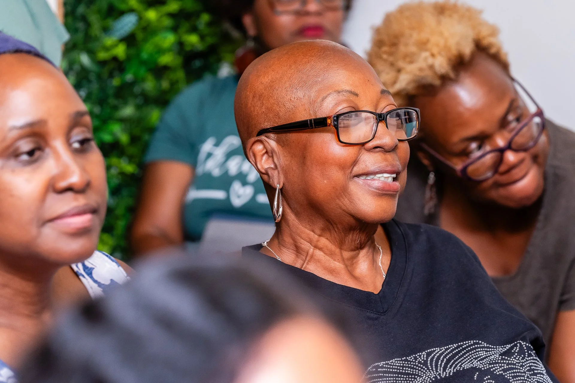 Close-up of three women sitting together, with the woman in the middle wearing glasses, a short hairstyle, and smiling, while the women on either side are partially visible.