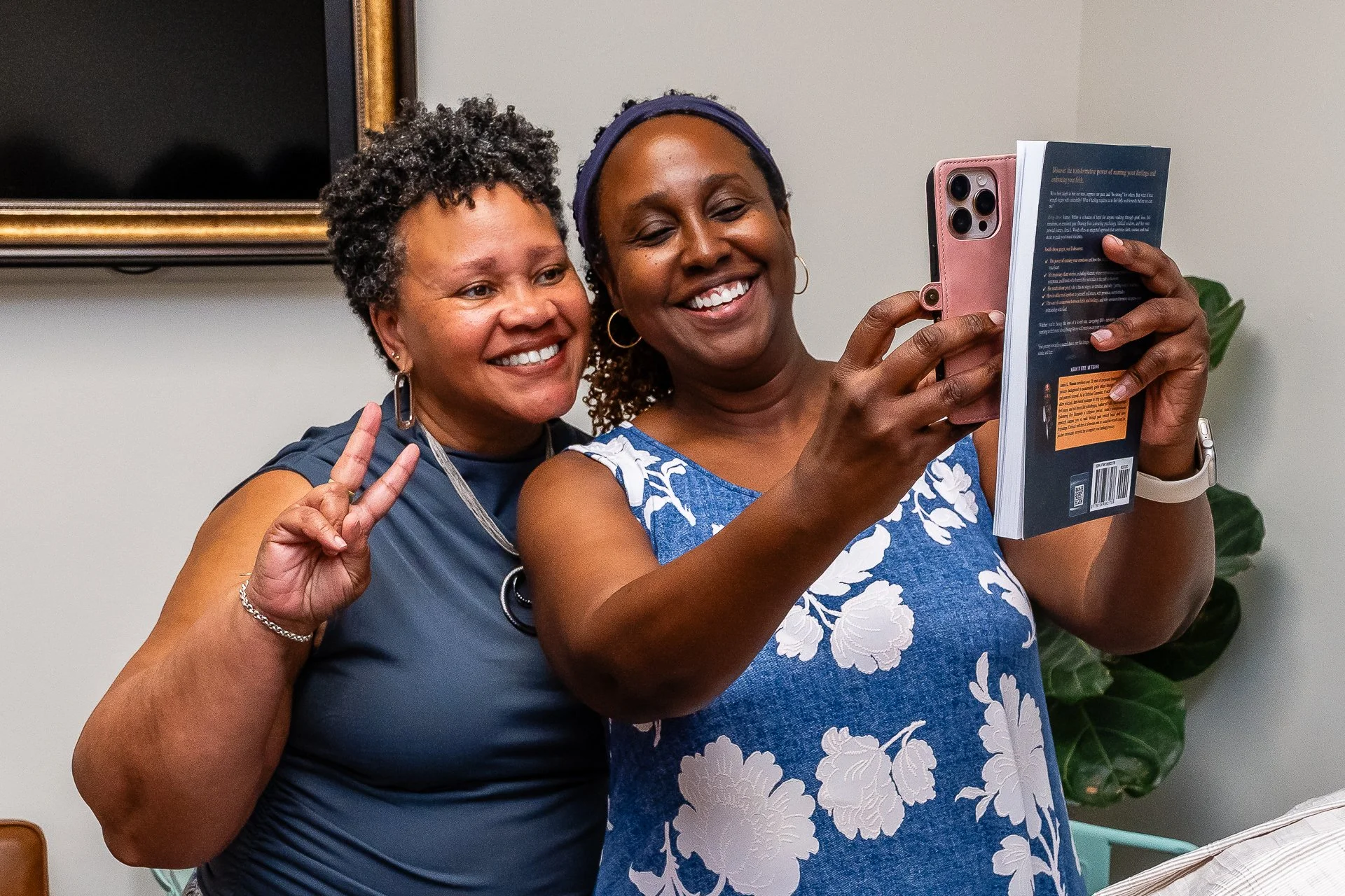 Two women taking a selfie together, smiling, with one making a peace sign. One woman is holding a book or magazine while the other has short curly hair and is wearing a dark sleeveless top. The setting appears to be indoors with a plain wall and a pl
