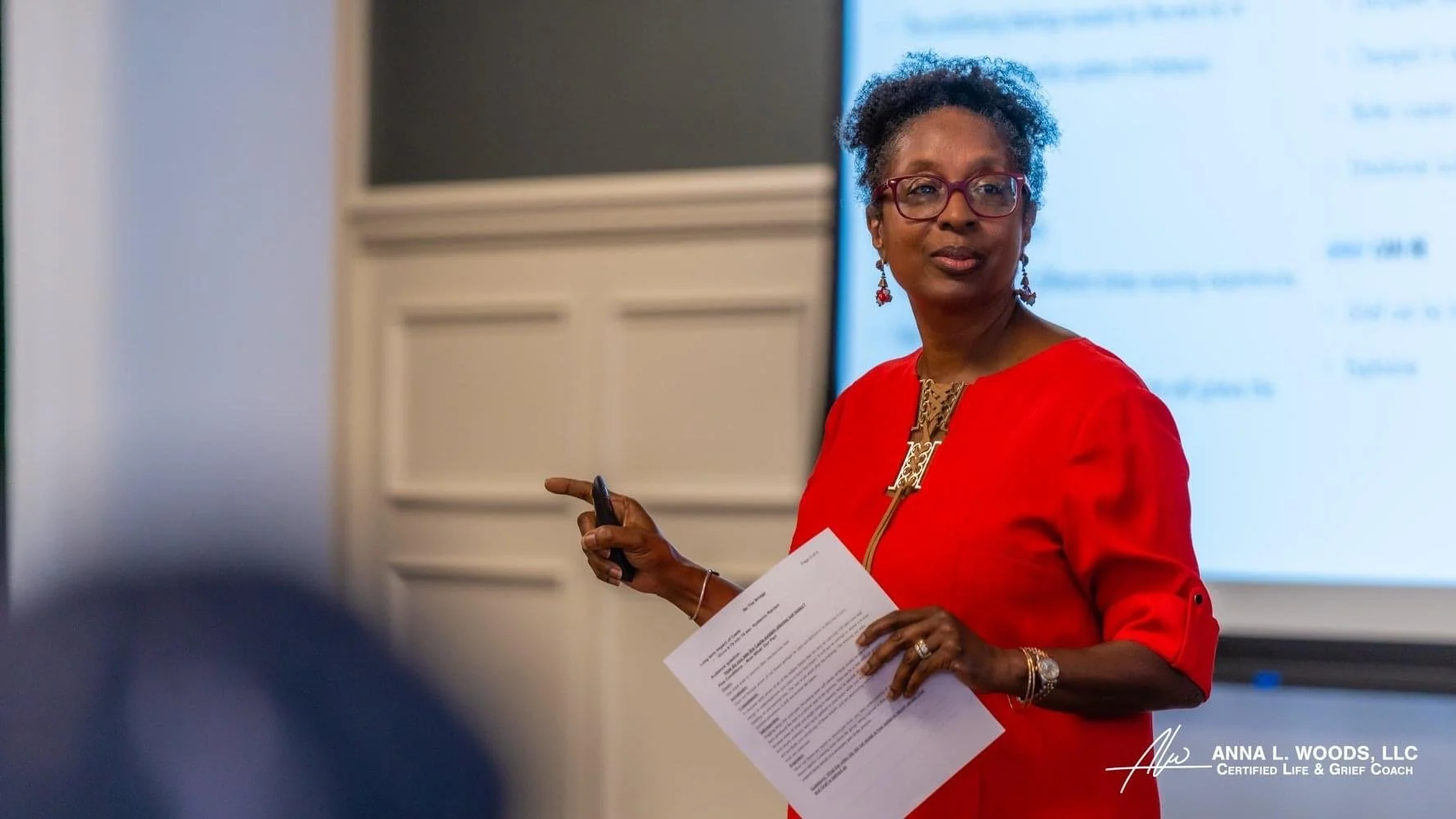 A woman with short curly hair and glasses wearing a red dress, holding a paper and a pointing device, appears to be giving a presentation in a classroom or conference room.