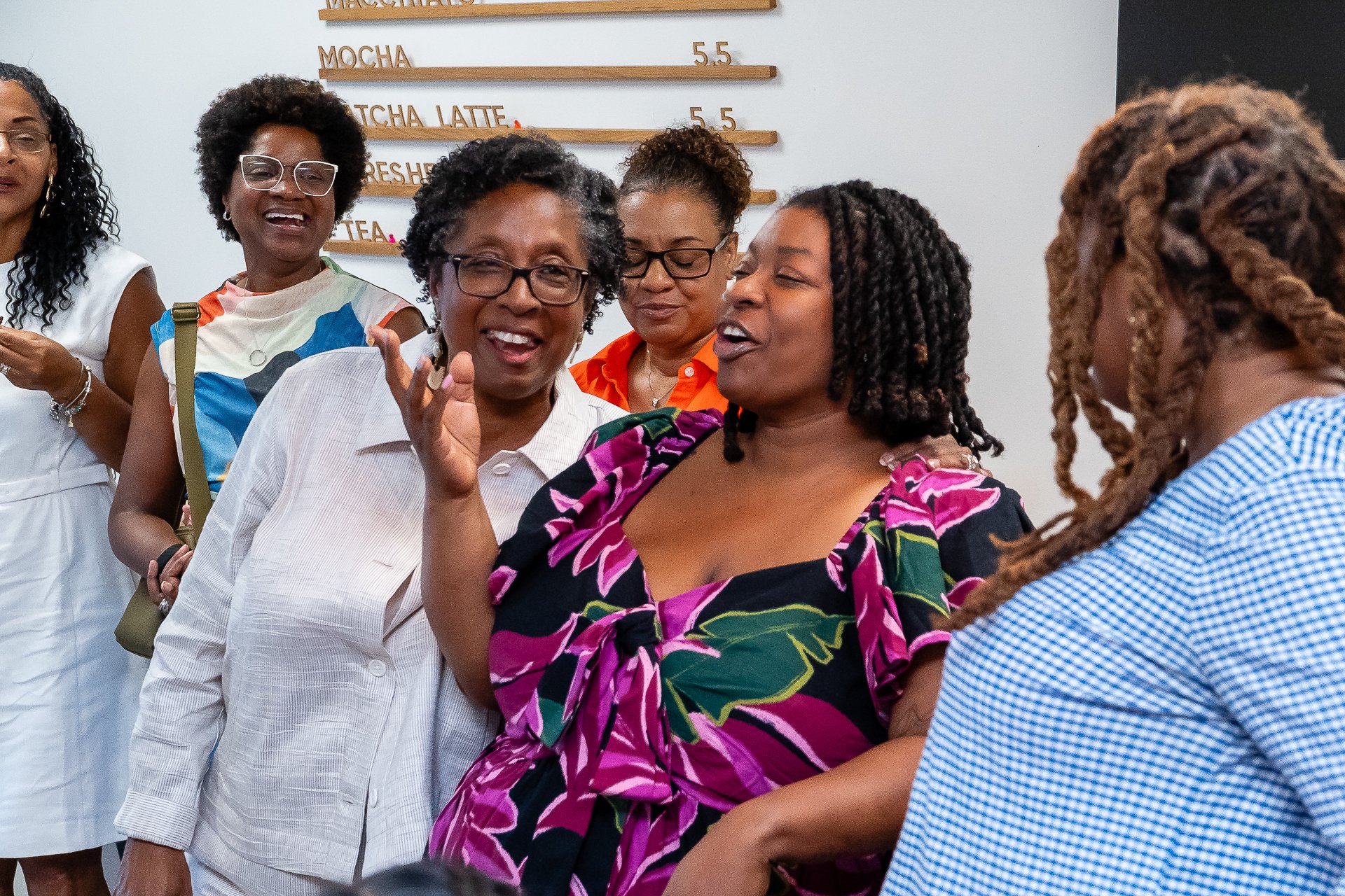 A group of diverse women engaged in conversation, smiling, and sharing laughter in an indoor setting, with a menu on the wall in the background.
