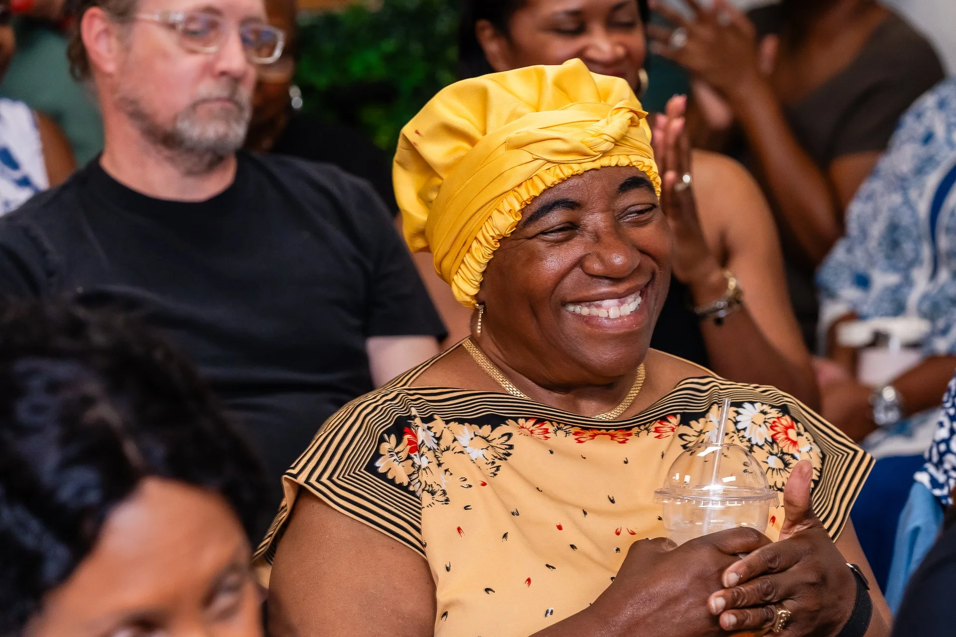 A woman wearing a yellow headwrap and a floral top sitting at a gathering, smiling and holding a plastic cup.
