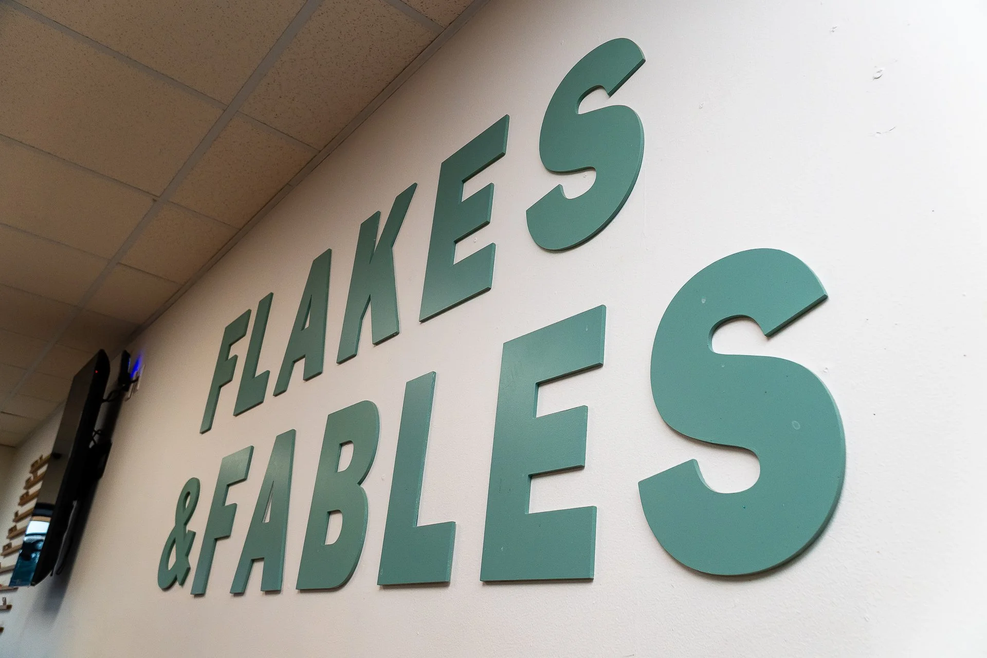 Close-up of large teal 3D letters on a white wall spelling out 'FLAKE 6', with a cereal store interior in the background.