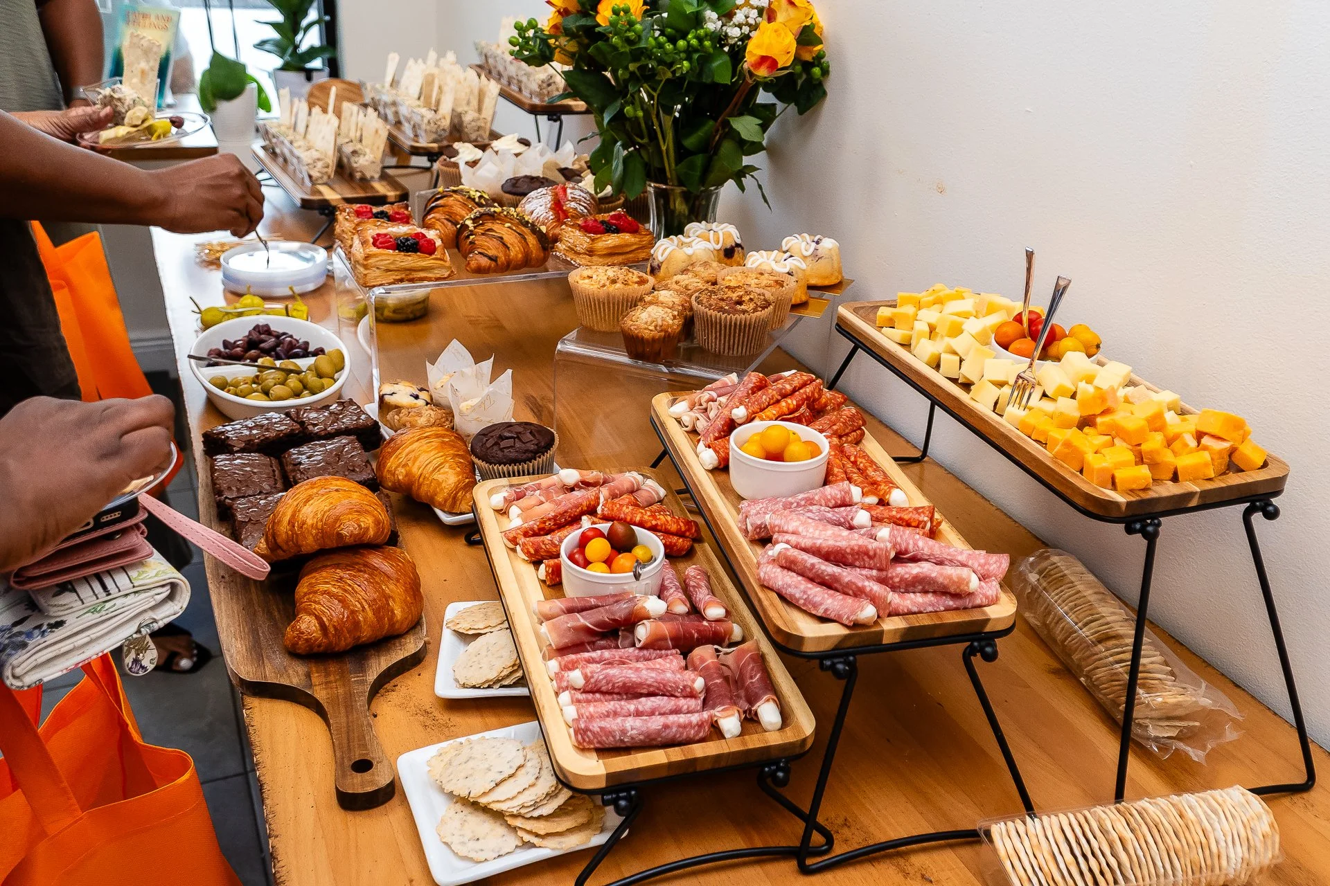 Buffet table with assorted baked goods including croissants, muffins, and pastries, along with cheese, cured meats, fruits, olives, and crackers, decorated with a large bouquet of flowers in a glass vase.