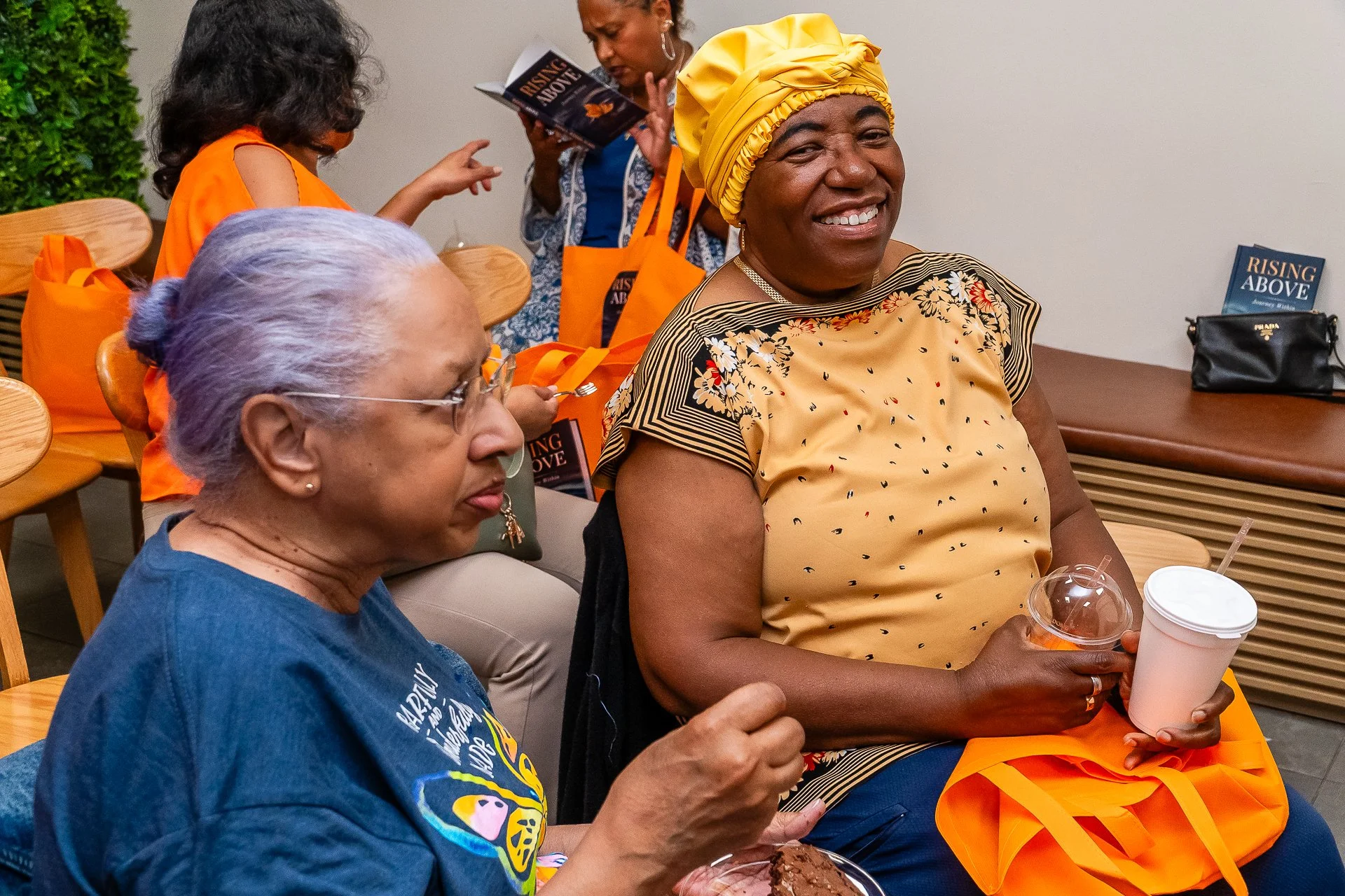 Smiling woman wearing a yellow headwrap and patterned shirt, holding a drink, sitting at a table with others during a gathering.