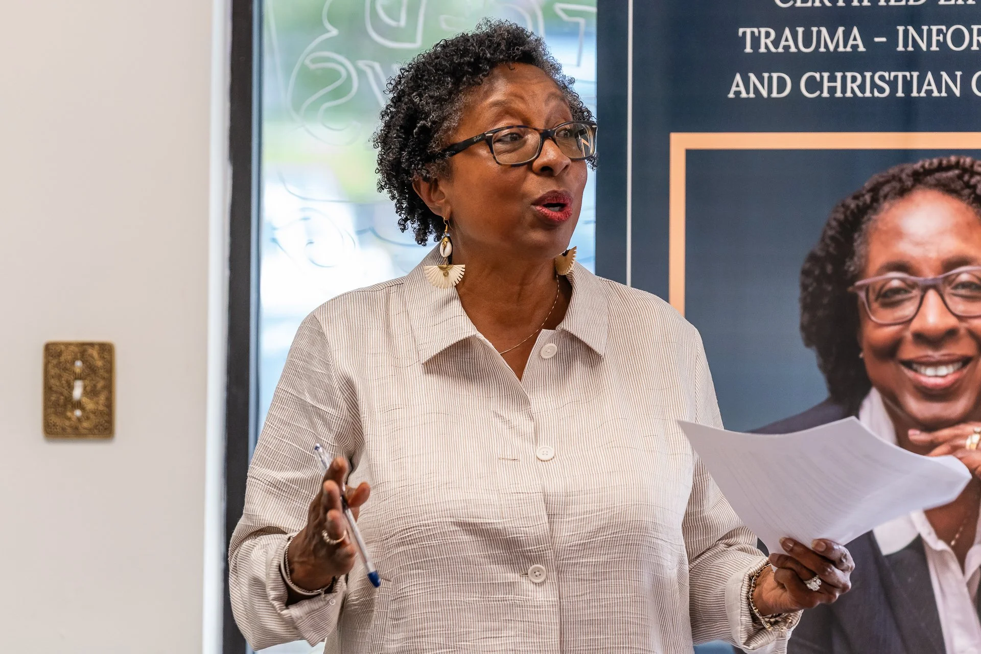 A woman with curly hair and glasses giving a presentation, holding papers and a pen, standing in front of a poster about trauma and Christian counseling, indoors near a light switch.