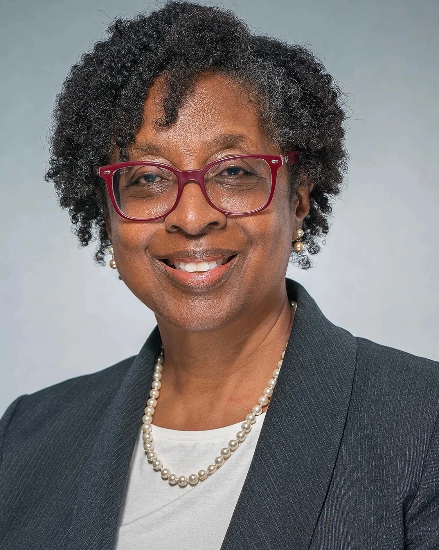 A professional portrait of an African American woman with curly salt-and-pepper hair, wearing red glasses, pearl earrings, a pearl necklace, a white top, and a dark blazer, smiling against a neutral background.