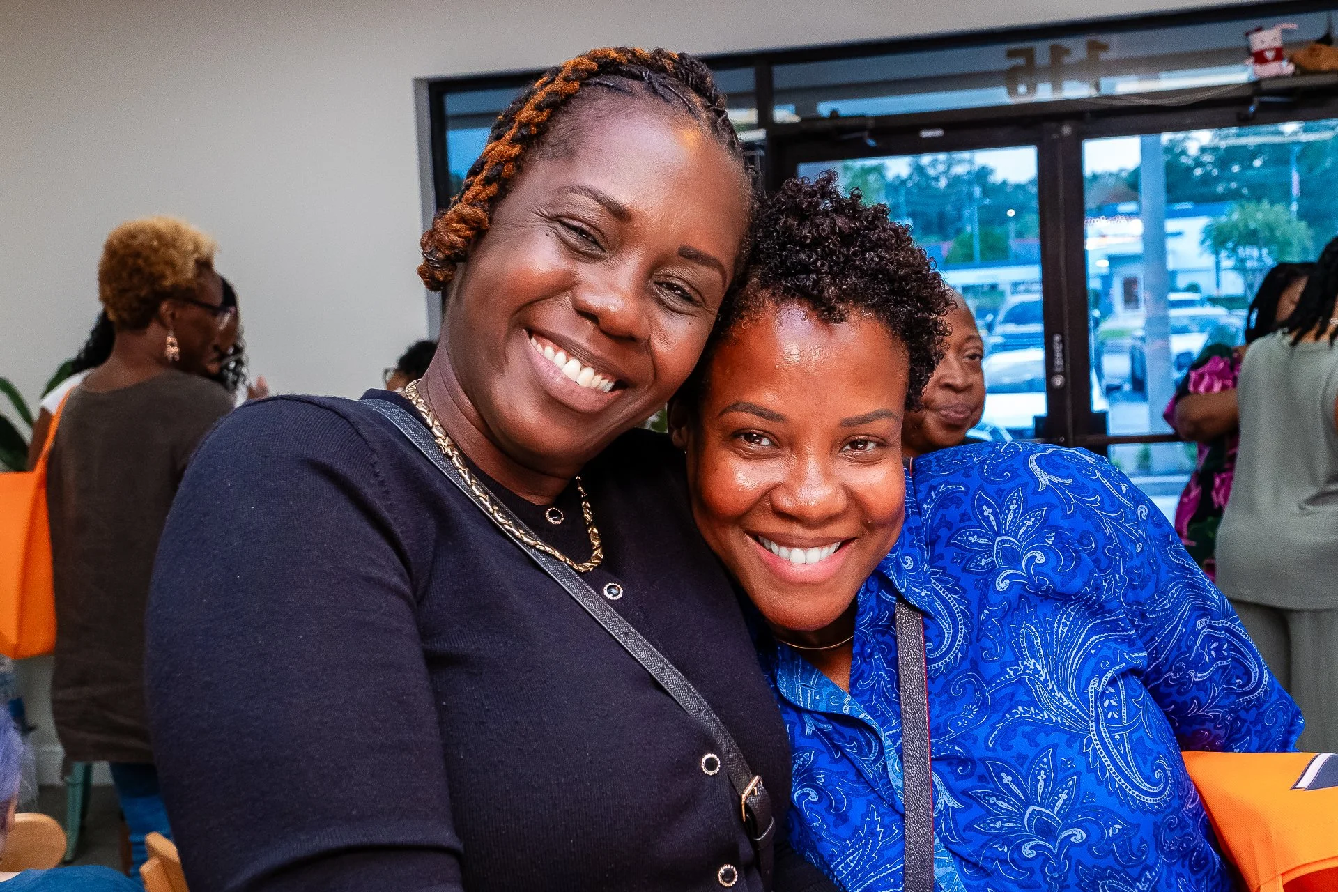 Two smiling women hugging in a social setting, with other people in the background.