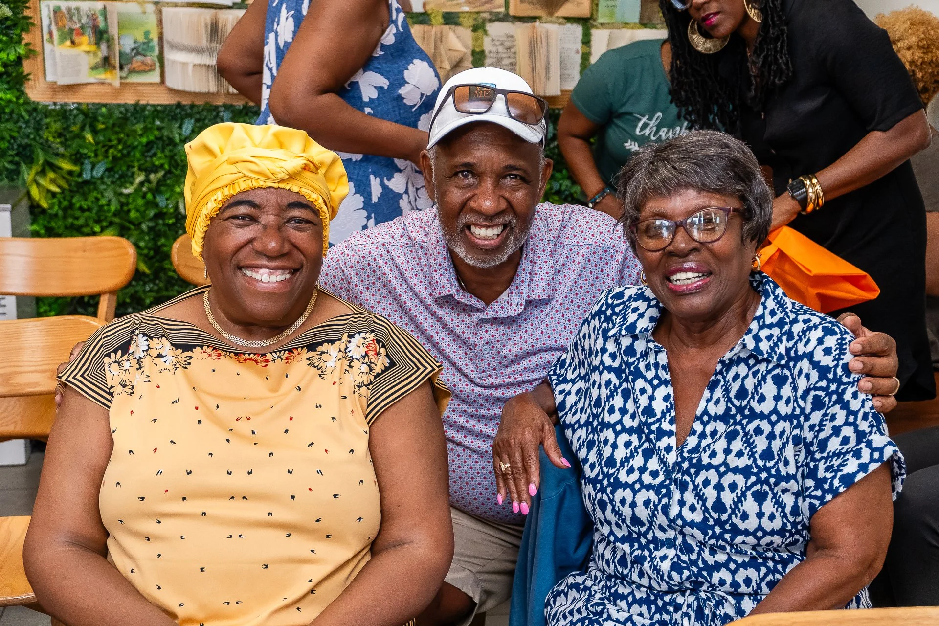 A group of diverse smiling older adults gathered in a cozy, well-decorated indoor space, sitting closely together at a table with wooden chairs, enjoying each other's company.