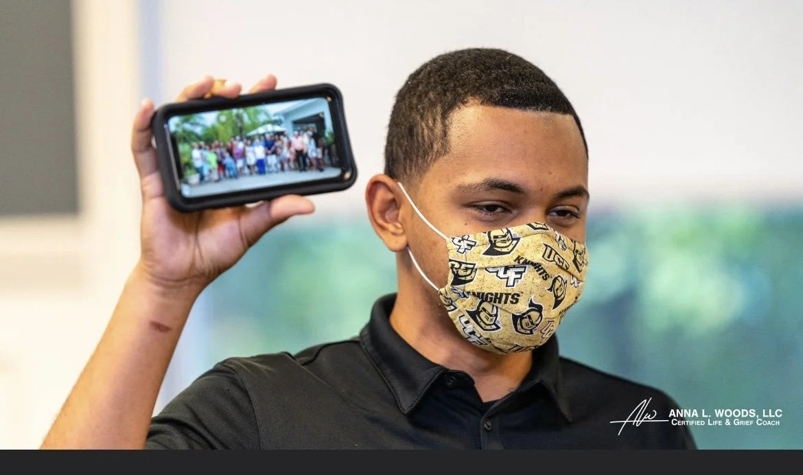 A young man wearing a face mask holding a smartphone displaying a group photo of people outdoors.