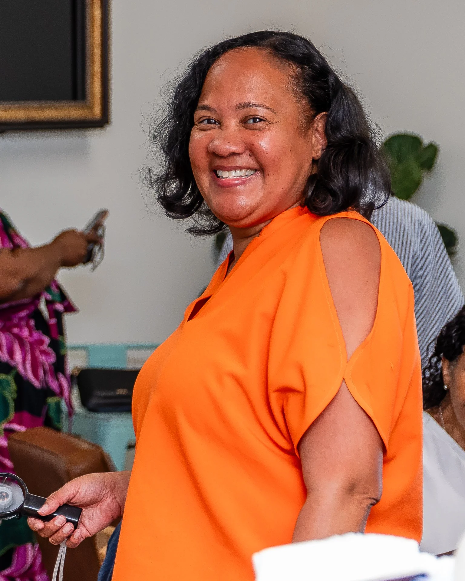 A woman smiling and wearing an orange top, holding an object in her left hand, with other people and green plants in the background.
