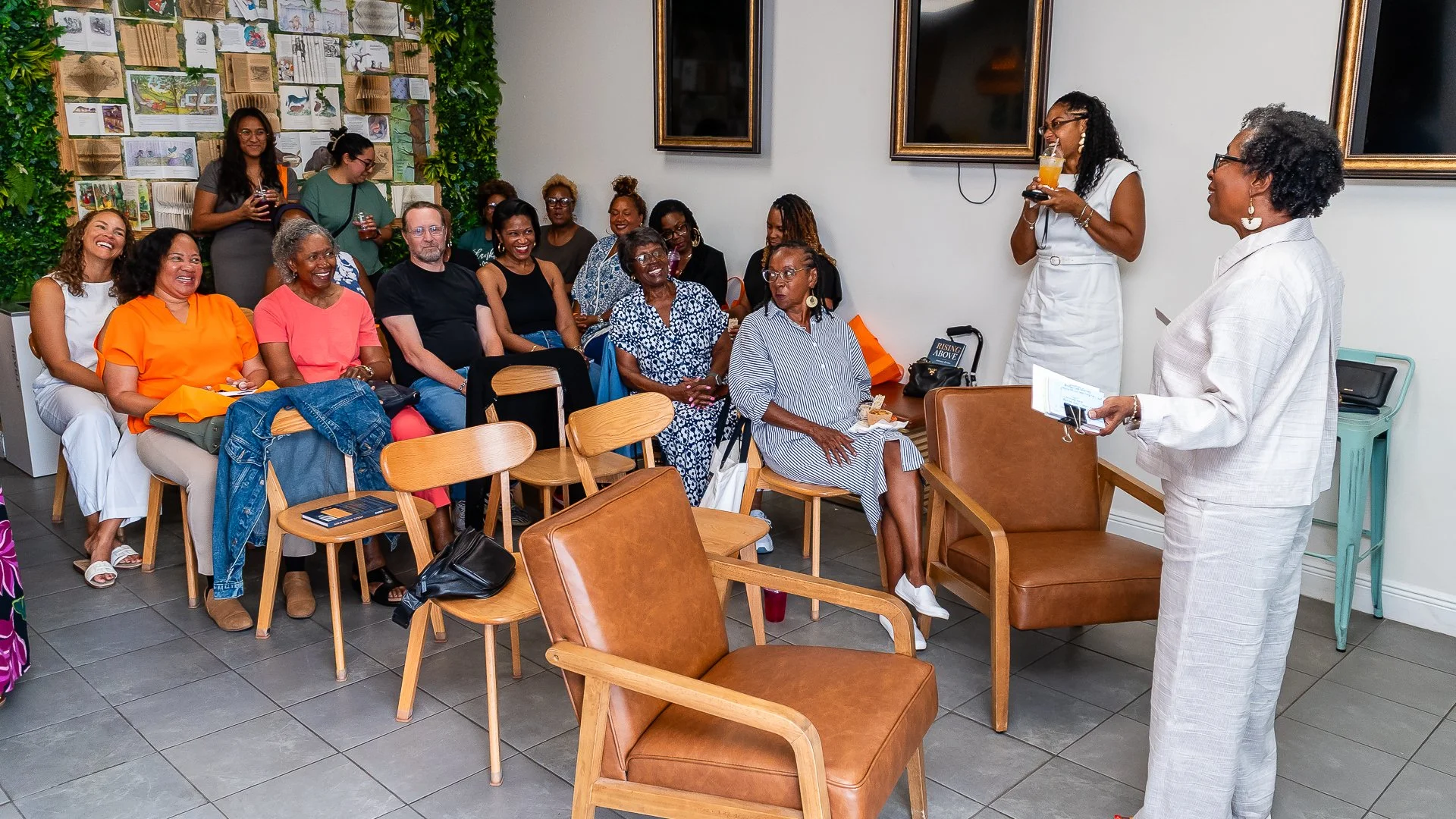 A group of mostly women sitting and smiling during a presentation or discussion in a room with a white wall, decorated with framed pictures and a green plant wall. Two women stand at the front, one speaking with a drink in hand, the other holding a p