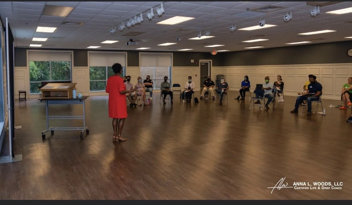 A woman in a red dress standing and speaking to a group of people sitting in chairs in a spacious room with large windows and wooden floors.