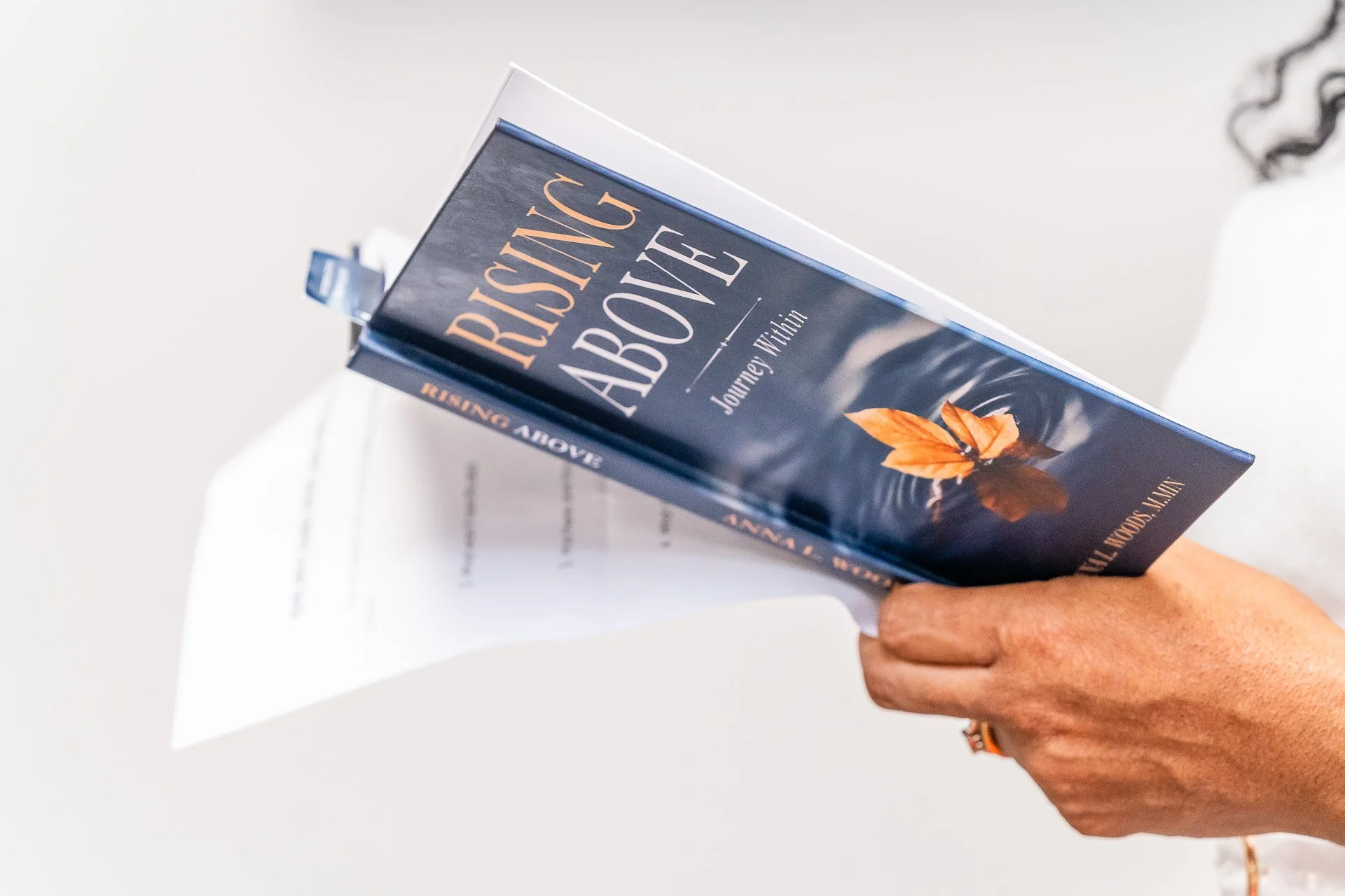 A person with tan skin holding a book titled "RISING ABOVE" by Jonney Withe, with a white piece of paper underneath the book. The person has curly hair partially visible on the right side of the image.