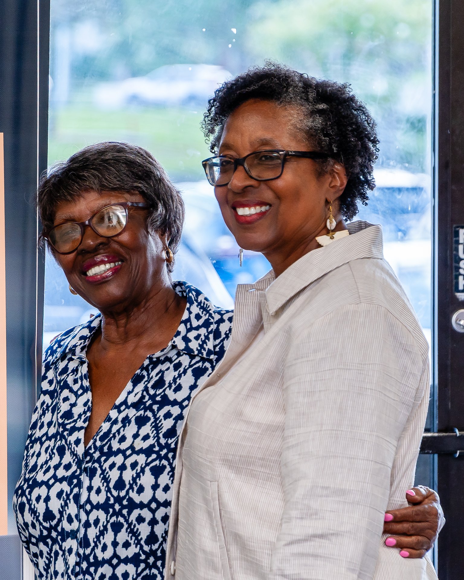 Two women smiling and embracing in front of a glass door with a blurred outdoor background.