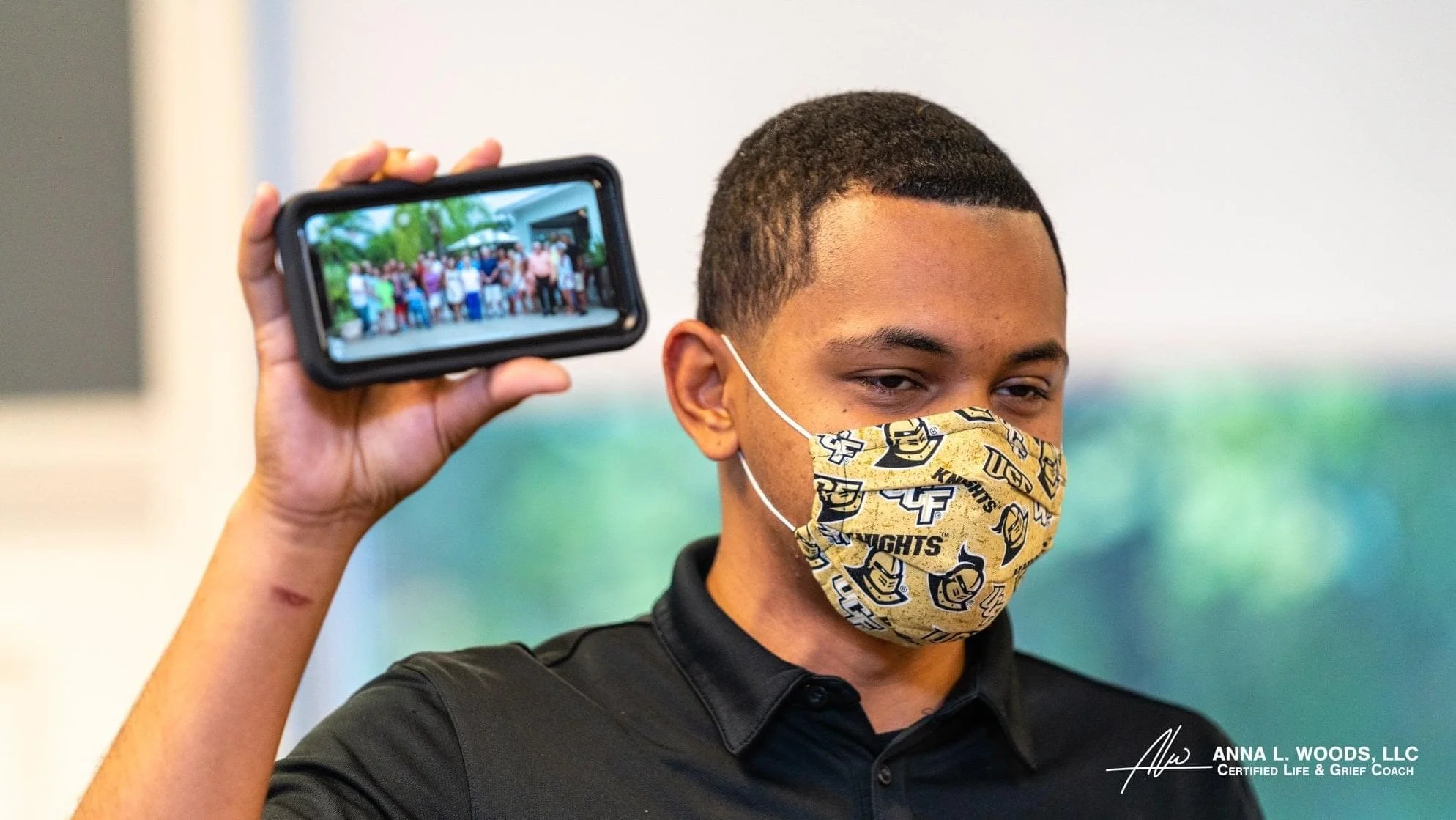 A young man in a patterned face mask holding a smartphone with a group photo displayed on the screen.