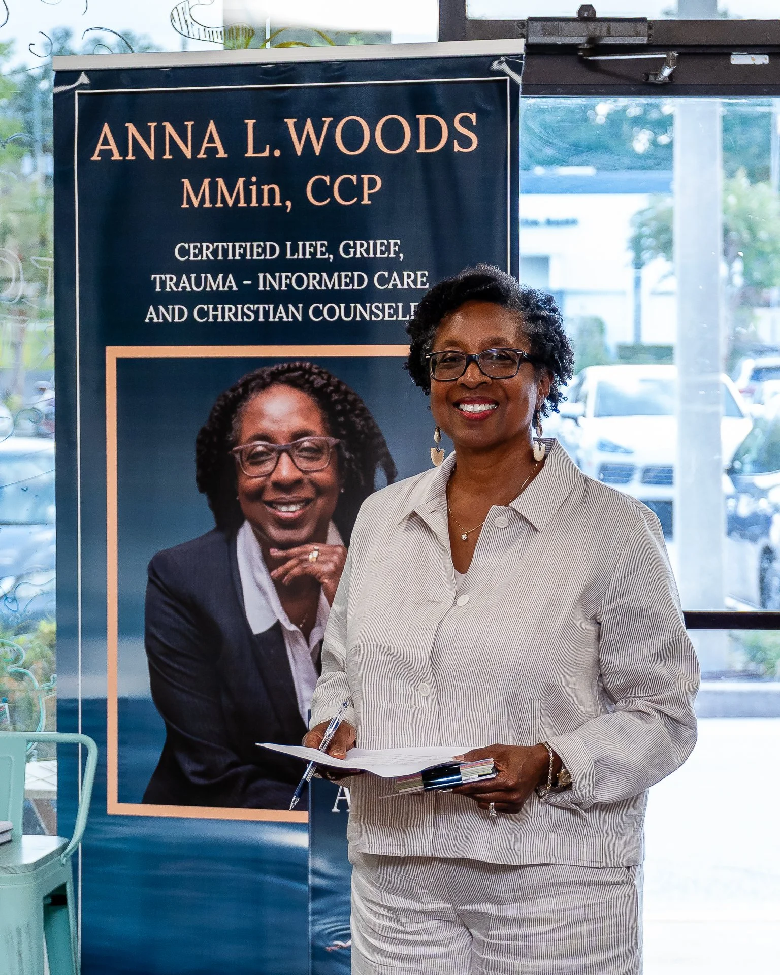 A smiling woman with glasses and short curly hair, holding a notebook and pen, standing in front of a sign that reads 'Anna L. Woods, MMIn, CCP, Certified Life, Grief, Trauma-Informed Care and Christian Counselor.'