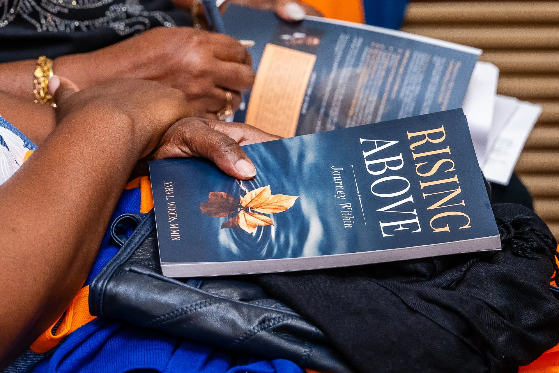Close-up of a person's hands holding a teal and orange paperback book titled 'Rising Above' by Journey With, resting on a navy blue bag with an orange strap, on a person's lap wearing a blue garment. The background shows a wood-paneled surface.