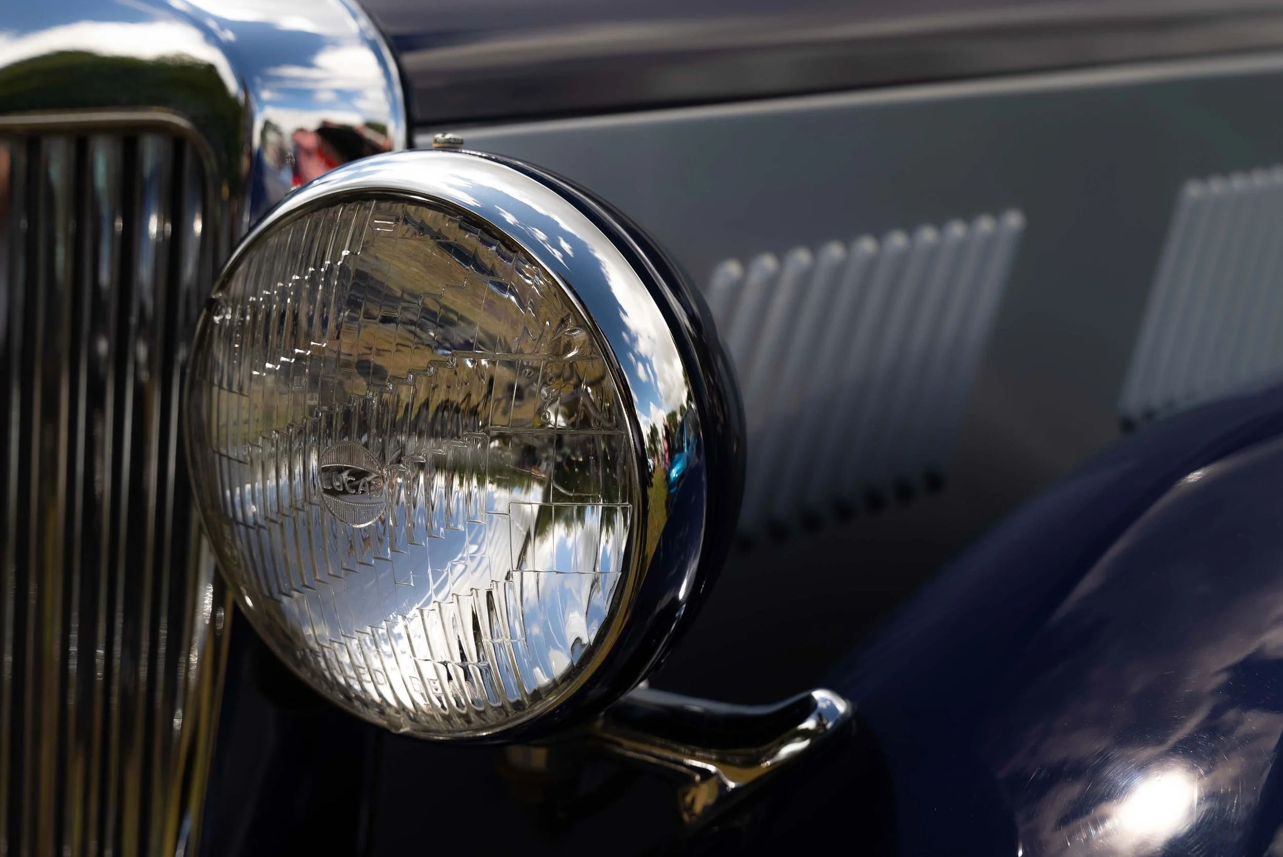 Close-up of a vintage car's headlight, grille, and body painted in dark color, with reflections of the sky and surroundings visible on the chrome surfaces.