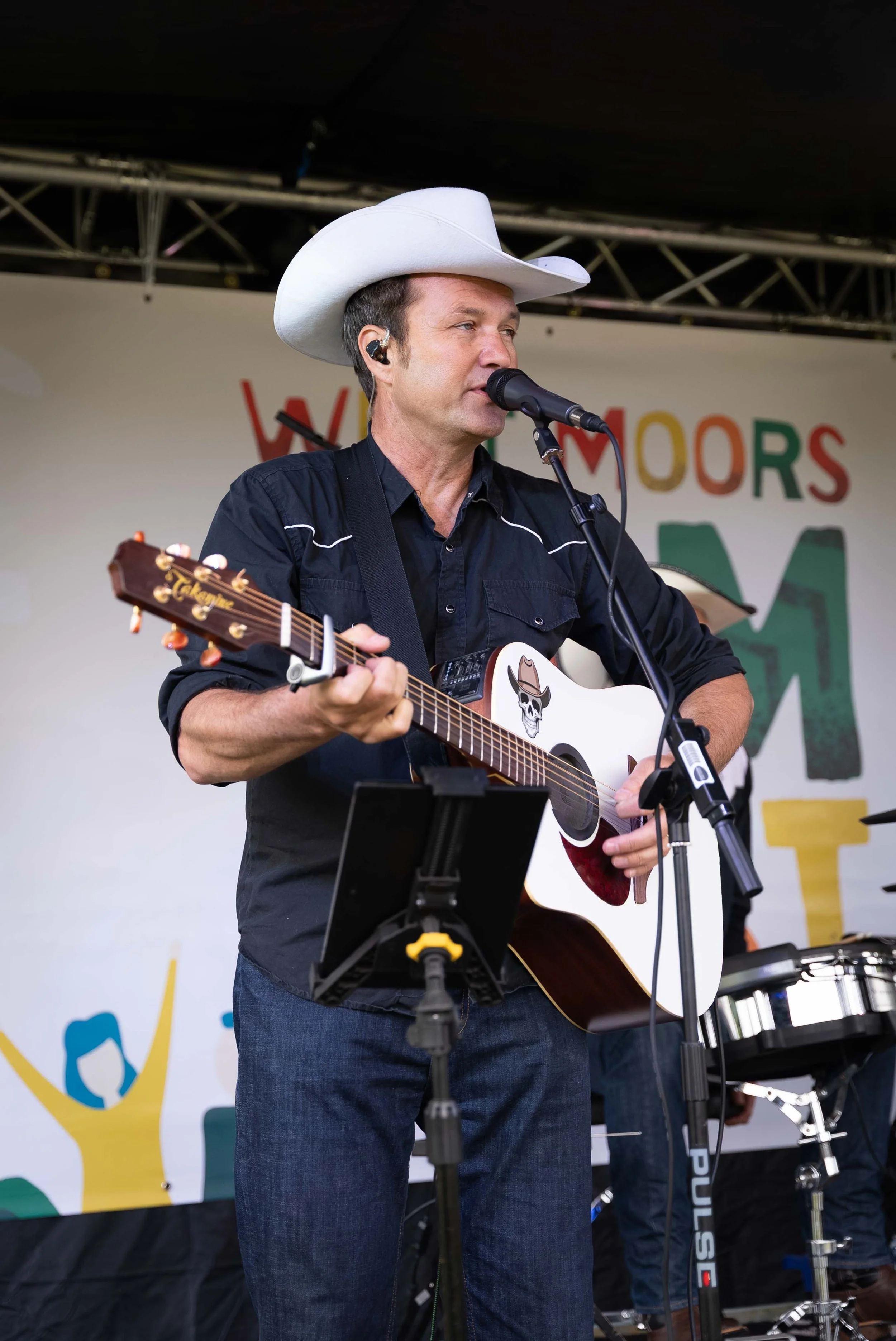 Male country musician singing into a microphone while playing an acoustic guitar at an outdoor event, wearing a white cowboy hat and black shirt.