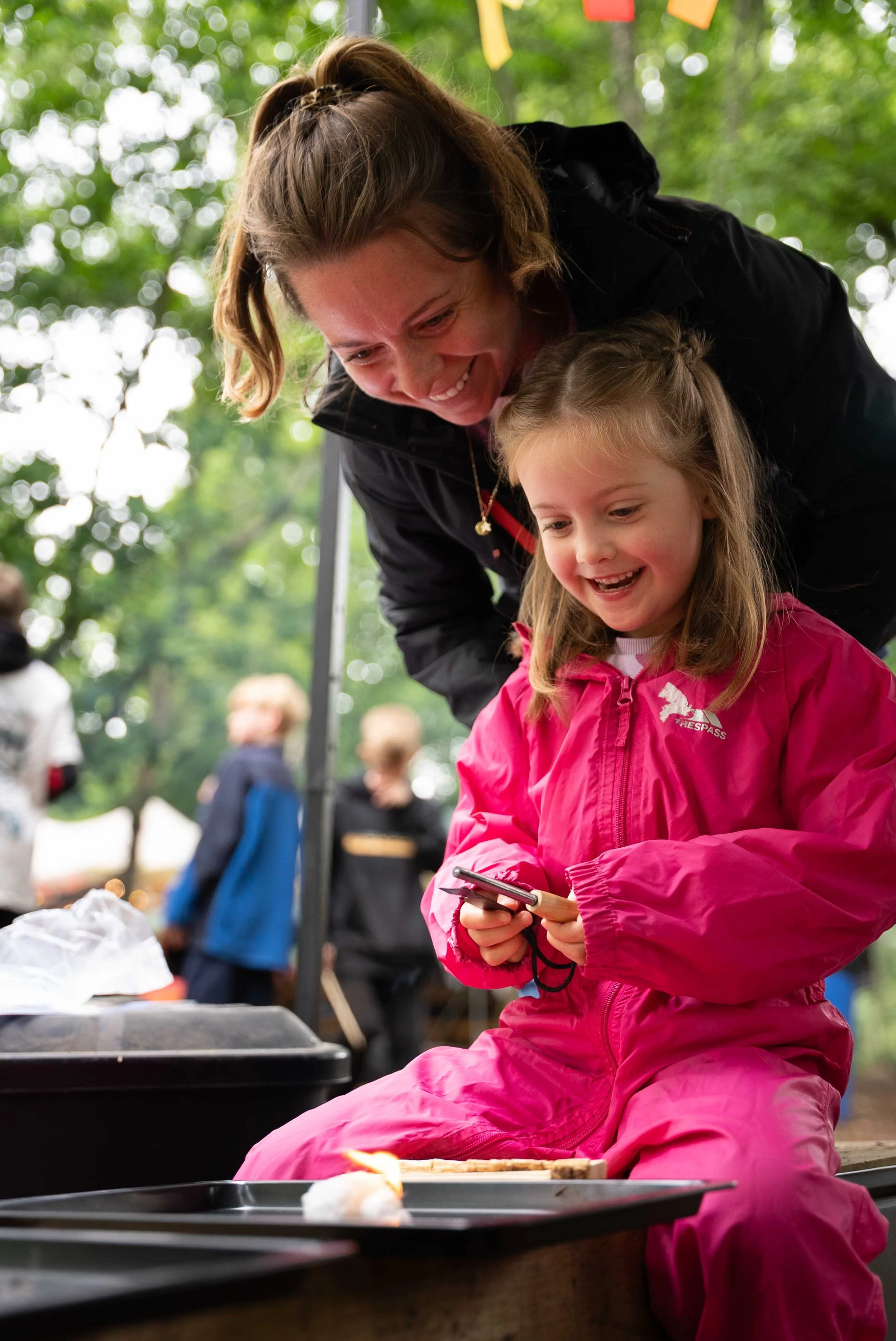 A woman and a young girl smiling and looking at a smartphone outdoors during a camping trip, with trees and other people in the background.