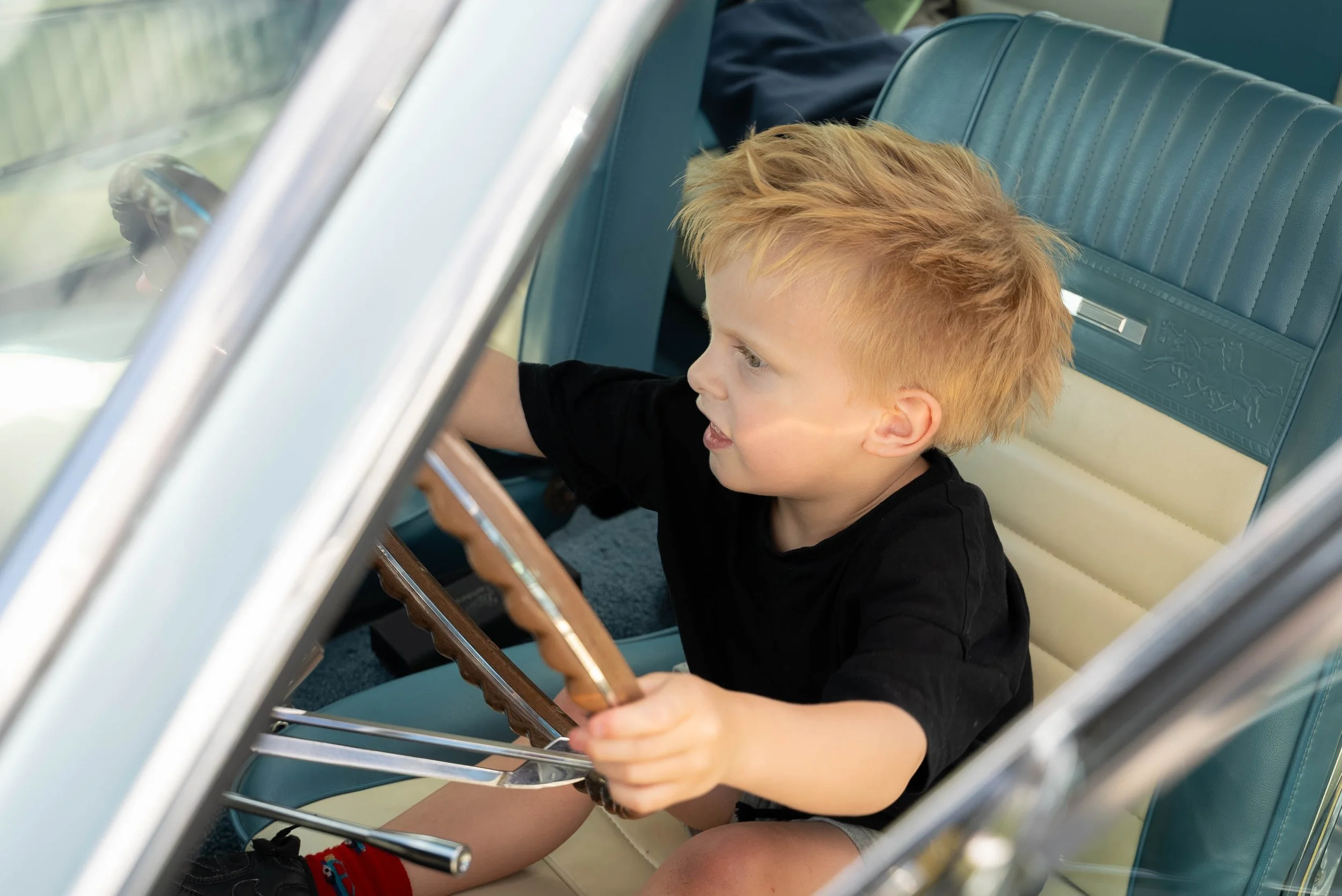 A young boy with blonde hair and a black shirt sitting in the driver's seat of a vintage car, holding the steering wheel and looking forward.