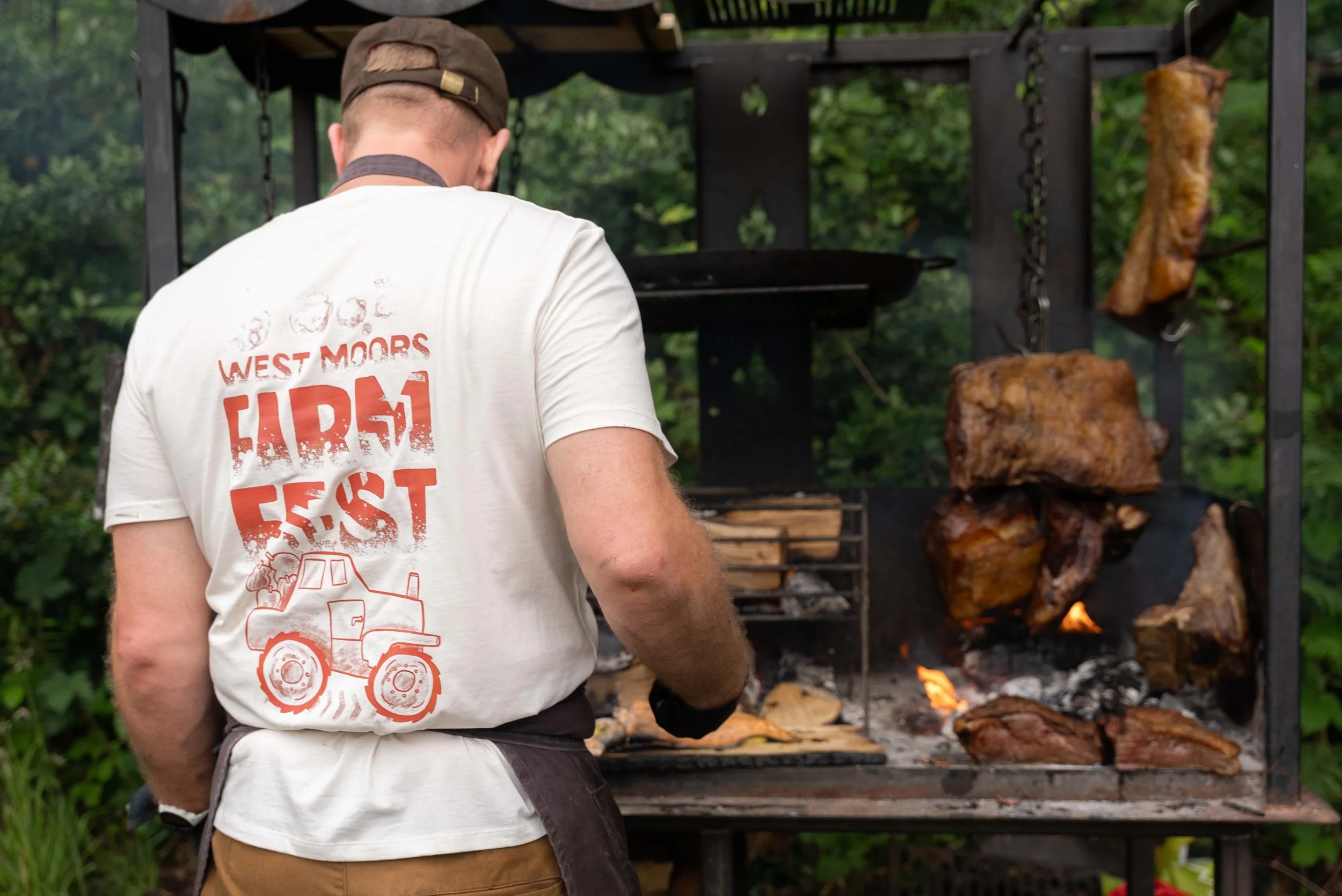 A man wearing a white t-shirt and a cap is grilling large pieces of meat over an open flame on a barbecue grill outdoors, with green foliage in the background.