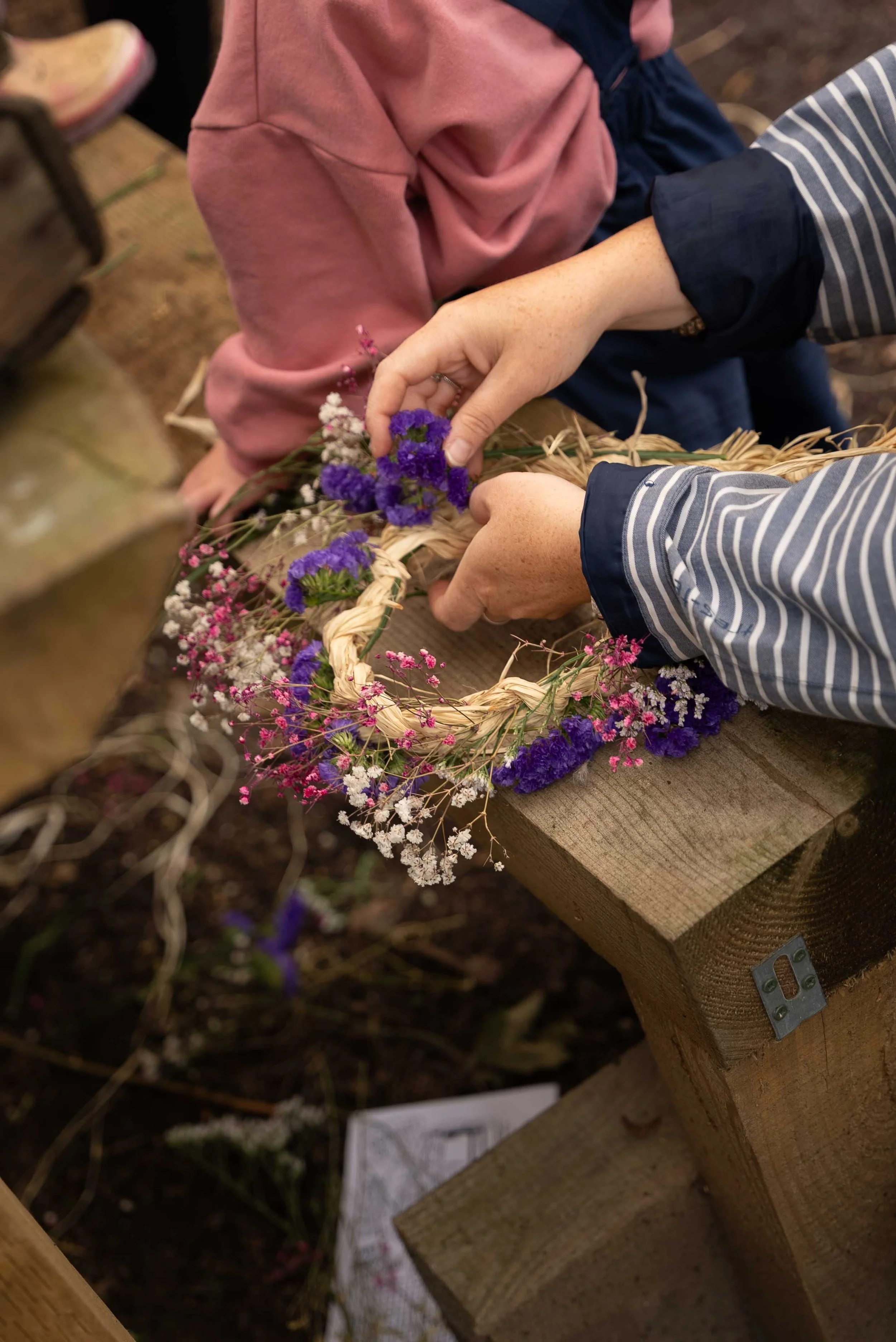 People are making a floral wreath with purple, pink, and white flowers, sitting outdoors on a wooden bench.