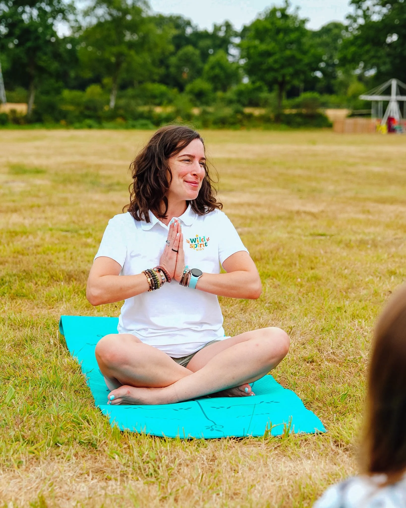 A woman practicing yoga outdoors on a blue mat in a park, sitting cross-legged with hands together in prayer position, eyes closed, smiling peacefully, wearing a white t-shirt with a colorful logo, surrounded by green trees and grass.